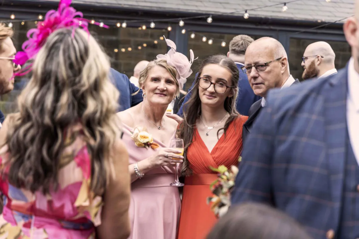Wedding guests mingling outdoors under string lights, with women wearing pink and orange dresses and stylish fascinators.