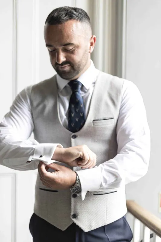 A groom in a light tweed waistcoat and patterned tie carefully fastens his cufflinks while getting ready on the morning of the wedding.