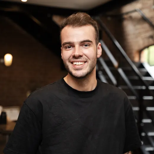 A smiling young man in a black t-shirt poses indoors against an industrial backdrop featuring exposed brickwork and dark metal beams.
