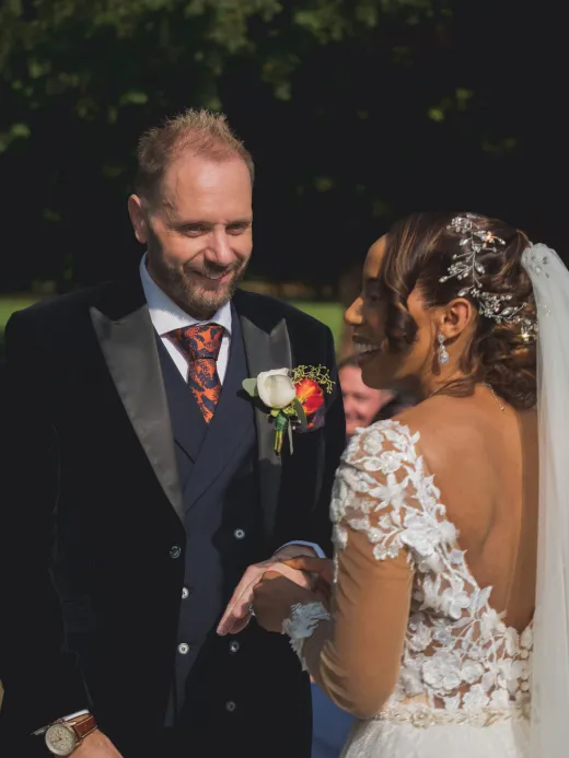 A joyful interracial couple share a tender moment holding hands during an outdoor wedding ceremony in natural sunlight.