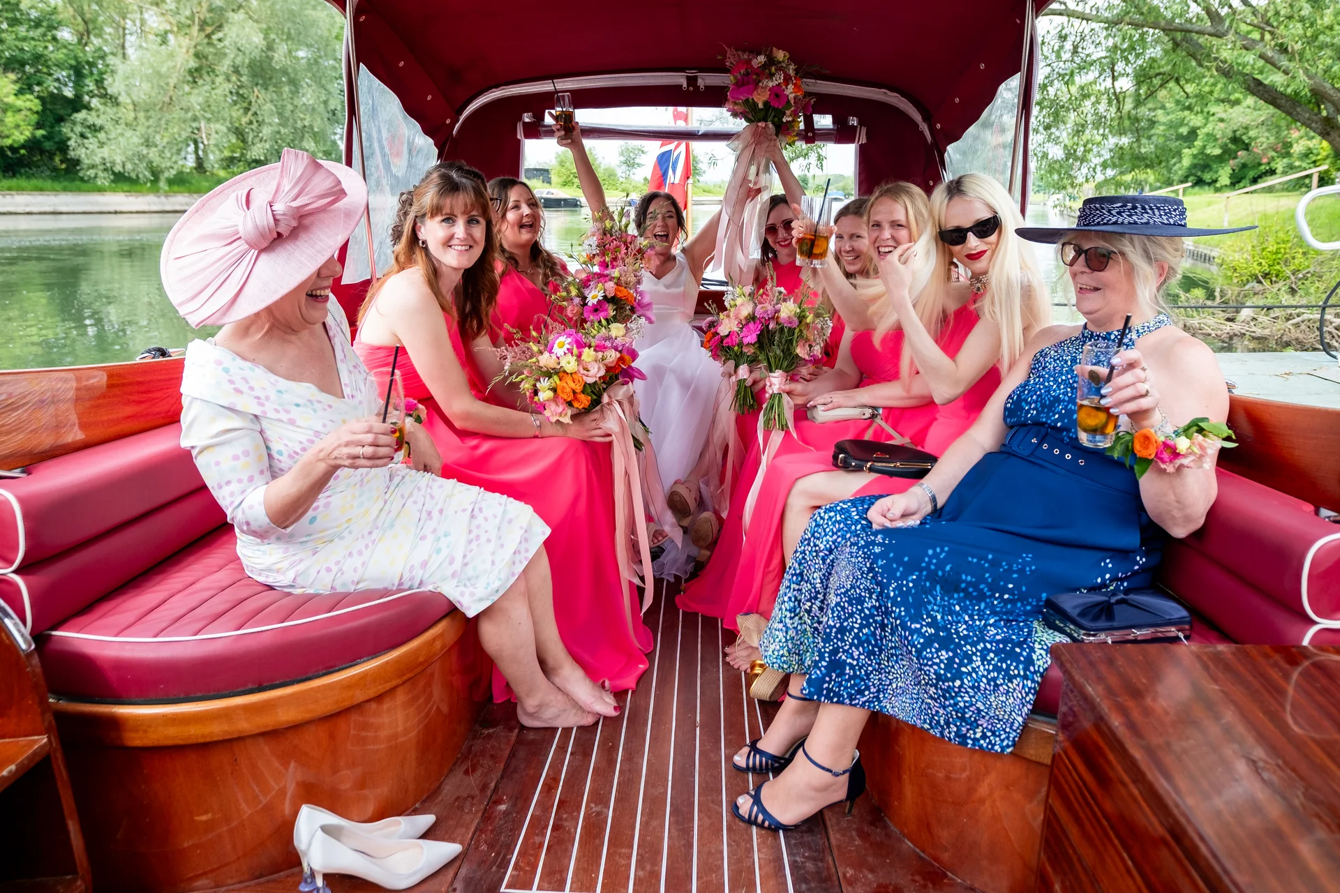 A joyful bridal party and guests celebrate with drinks inside a wooden boat on a sunny river during a wedding celebration.