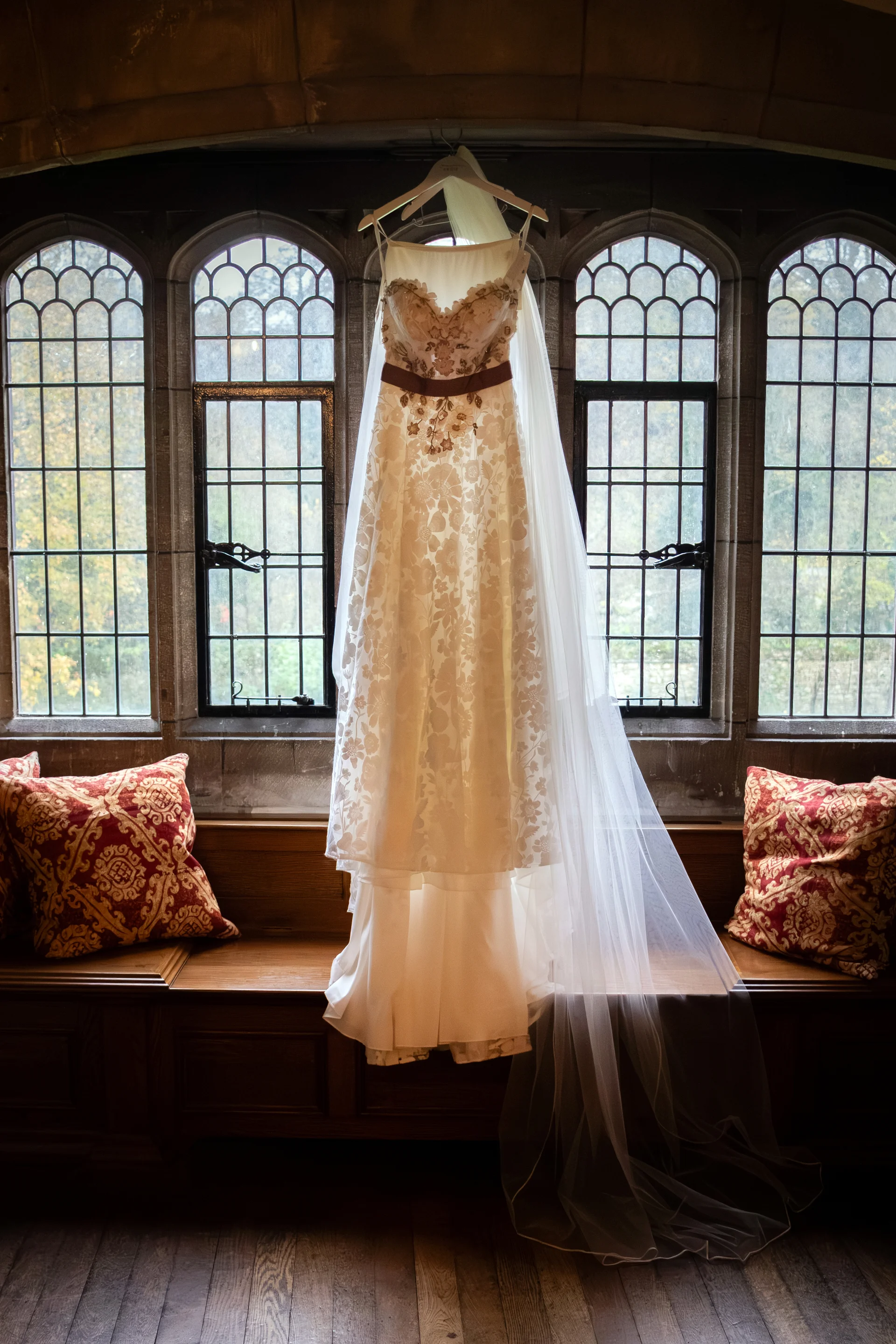 A detailed lace wedding dress hangs gracefully in front of large leaded windows, illuminated by soft natural light above a wooden bench with patterned cushions.