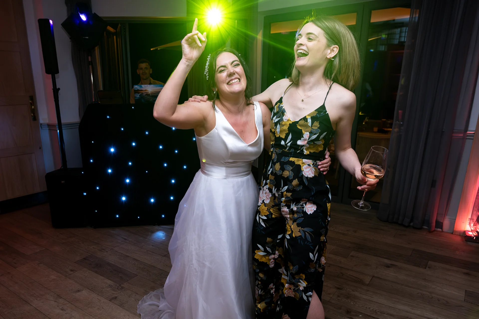 A joyful bride in a white gown and a guest dancing enthusiastically on the dance floor under bright green lighting near a DJ booth.