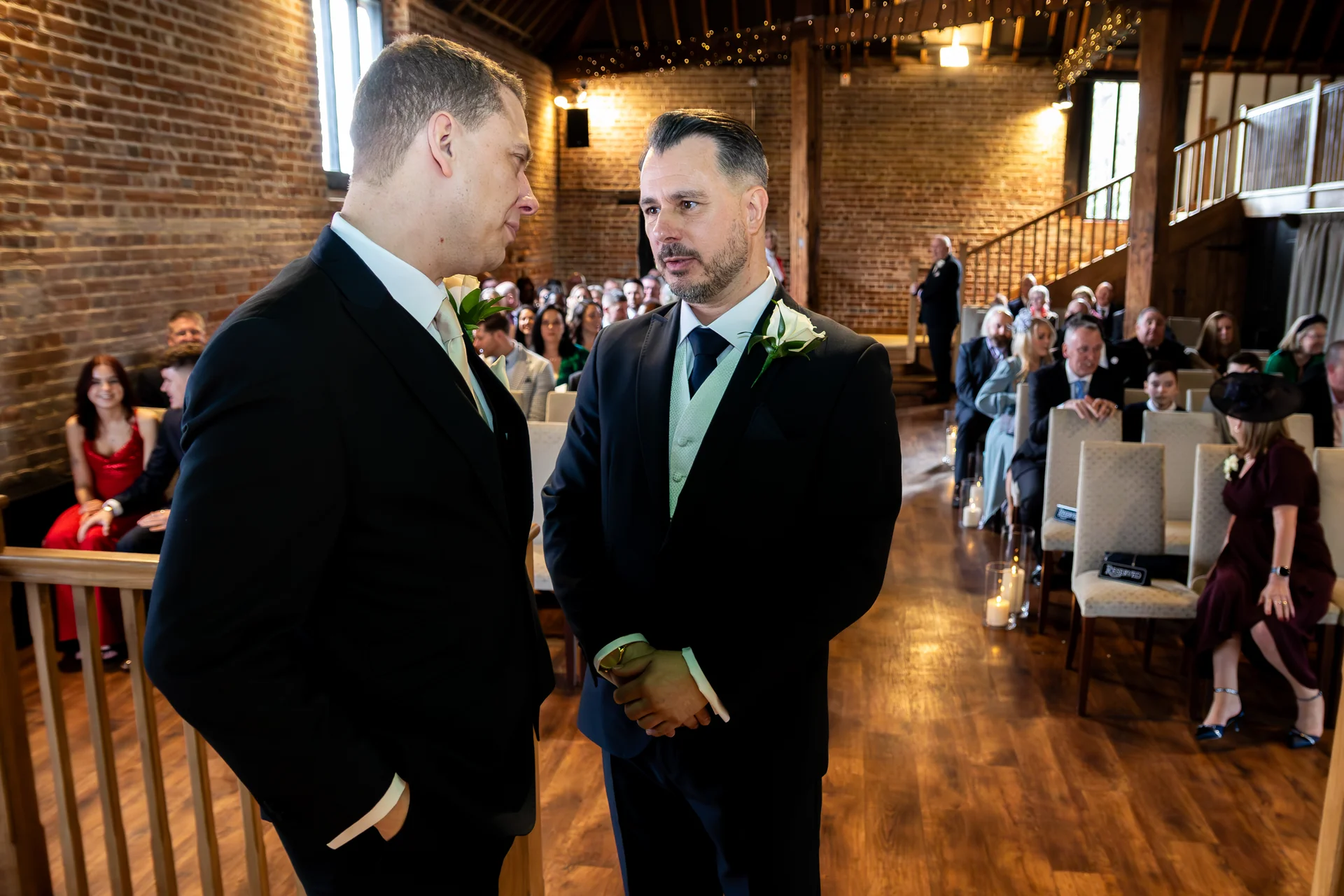 Two grooms stand together in a rustic brick barn venue during their wedding ceremony, surrounded by seated guests and wooden interiors.