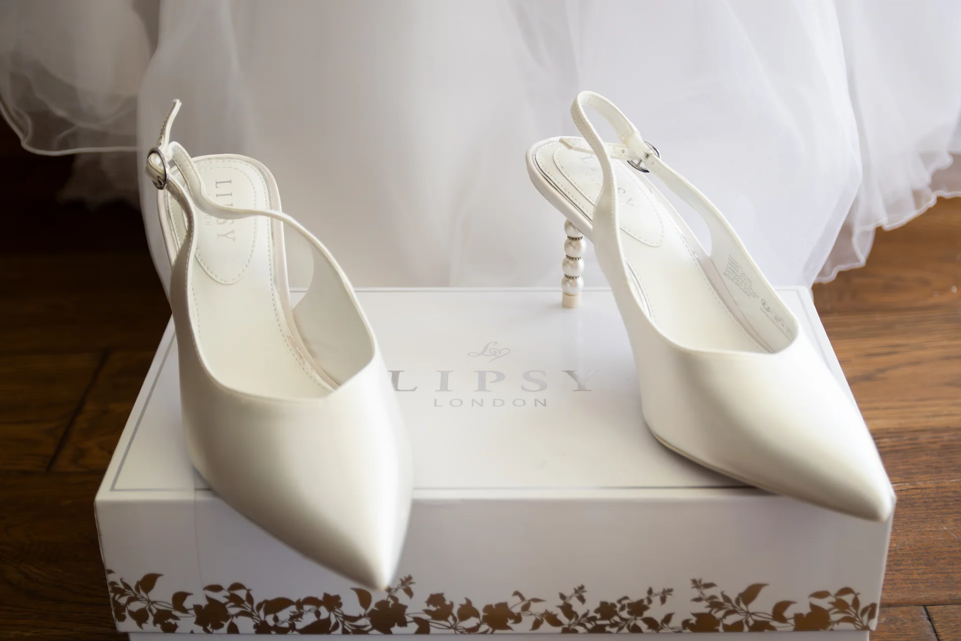 A pair of white satin slingback bridal shoes with pearl-embellished heels rests on top of their branded presentation box against a soft white dress.