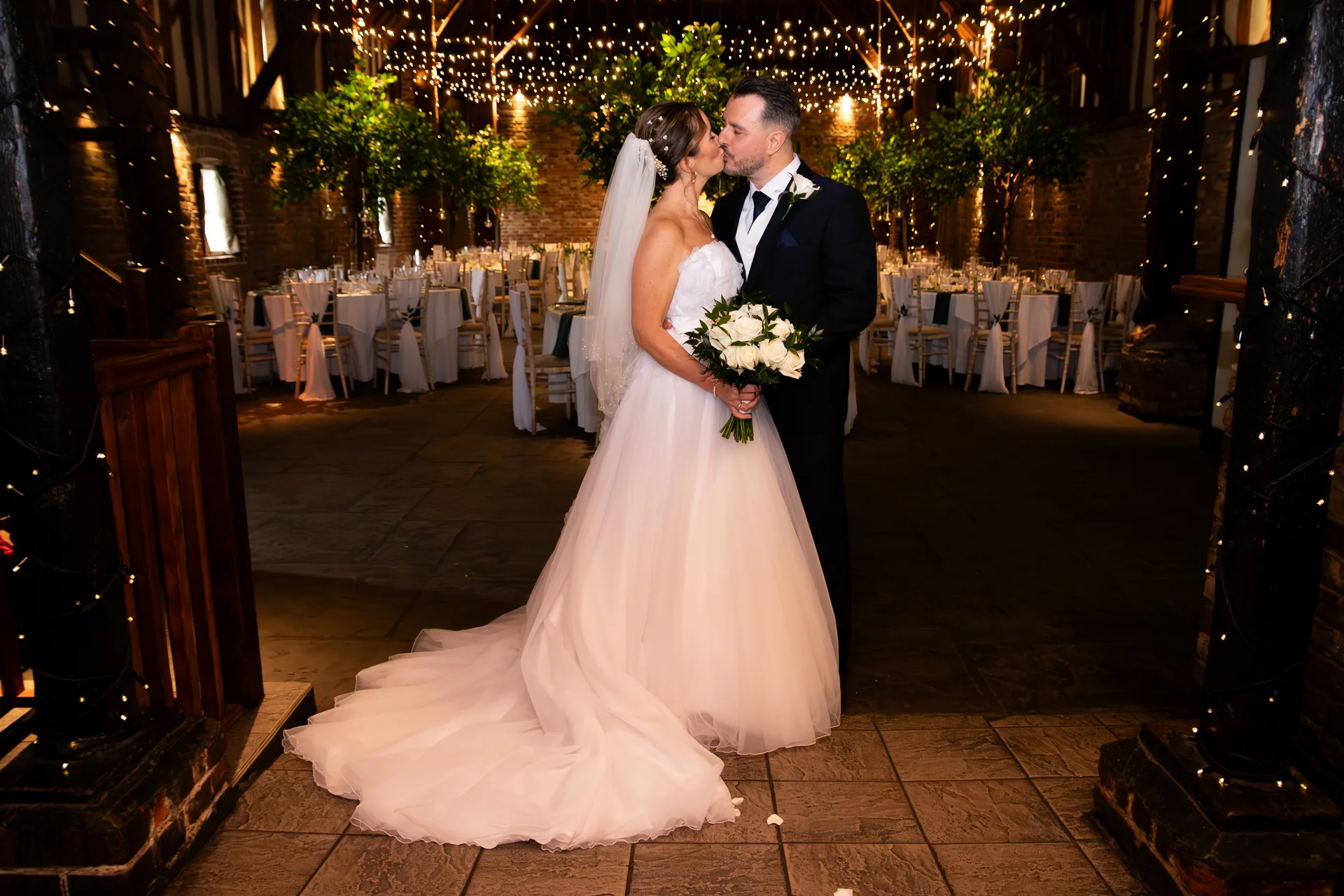A newlywed couple shares a tender kiss inside a rustic barn venue illuminated by a canopy of fairy lights and decorated tables.