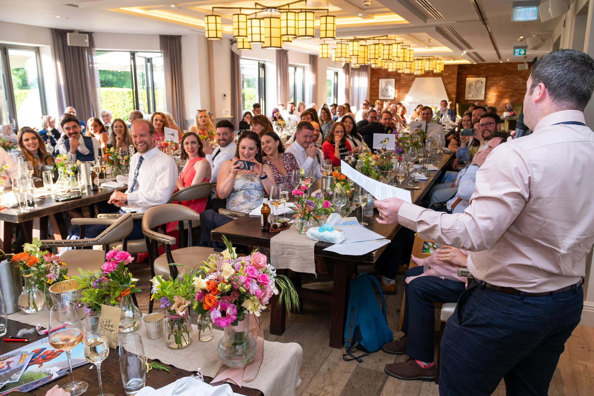 A guest delivers a speech holding notes at a wedding reception while seated guests laugh and listen in a brightly lit dining room.