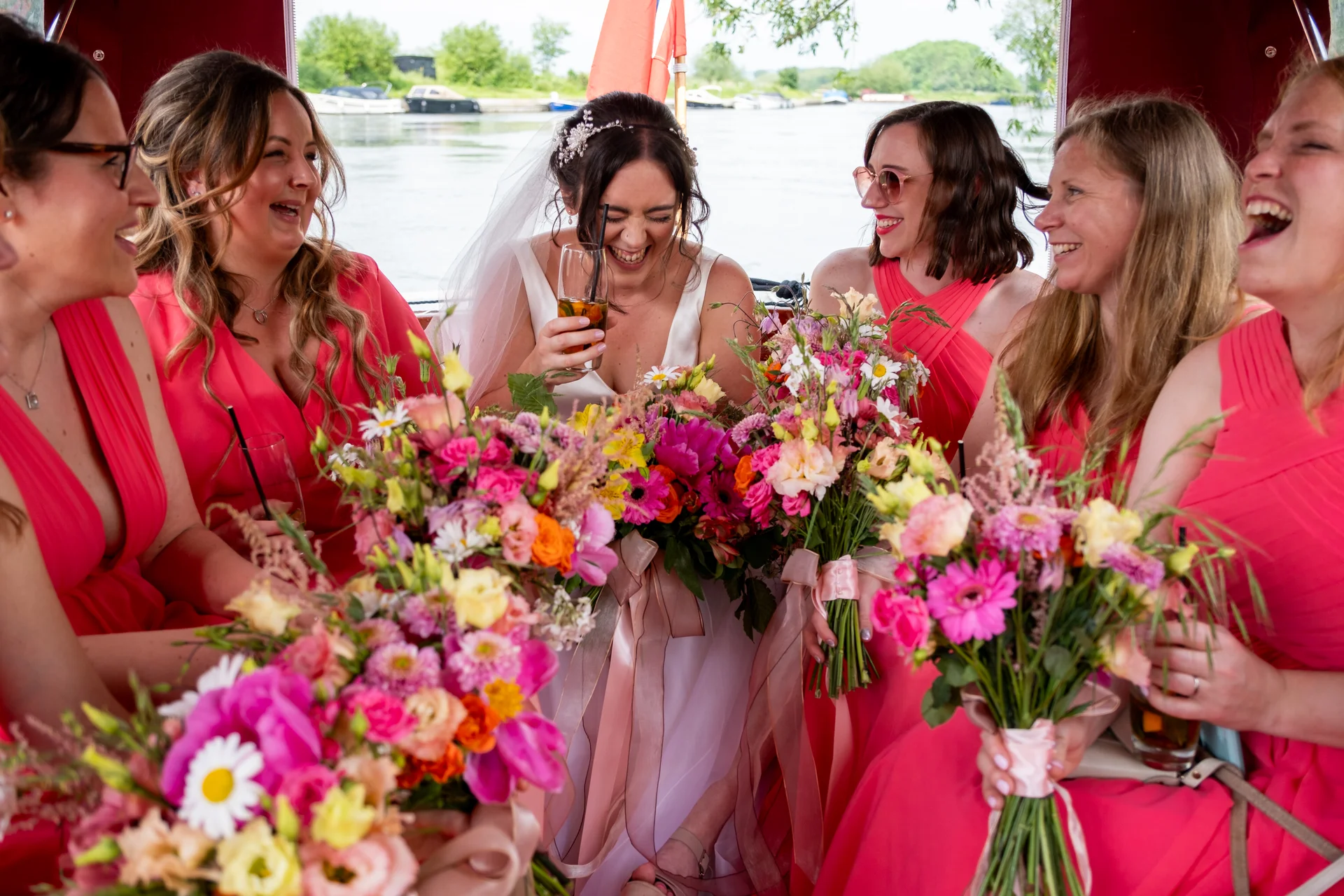 A joyous bride in a white gown and veil sits on a boat surrounded by laughing bridesmaids in bright pink dresses holding colourful floral bouquets.
