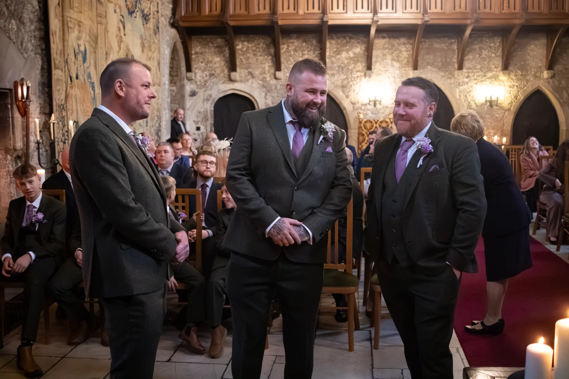 Three smiling groomsmen wearing matching tweed suits and purple ties stand inside a historic stone castle venue with guests seated in the background.