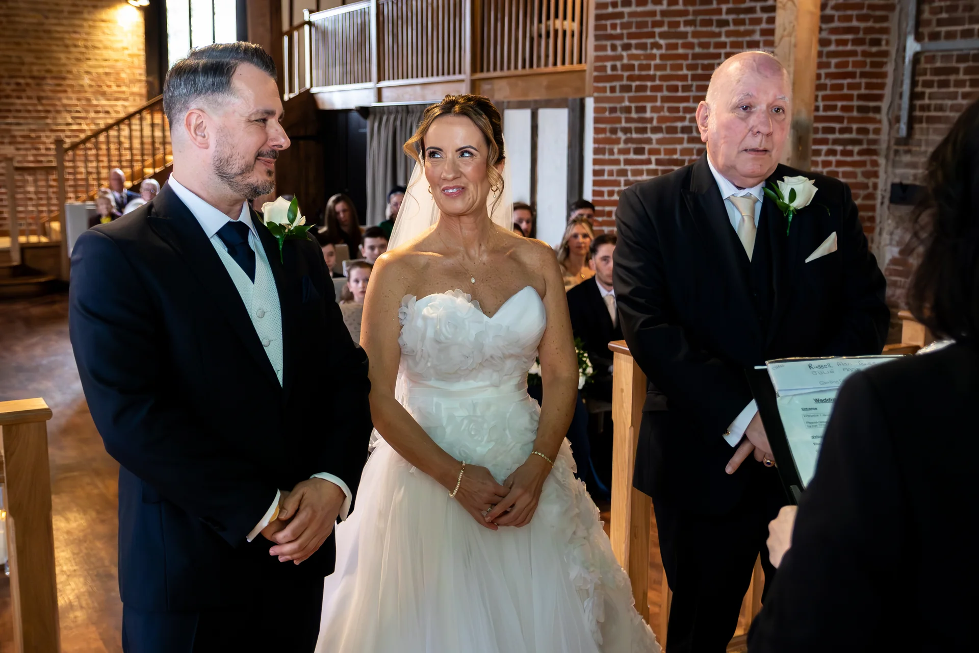 The bride and groom stand smiling at the altar inside a rustic barn venue with exposed brick walls, accompanied by the father of the bride.