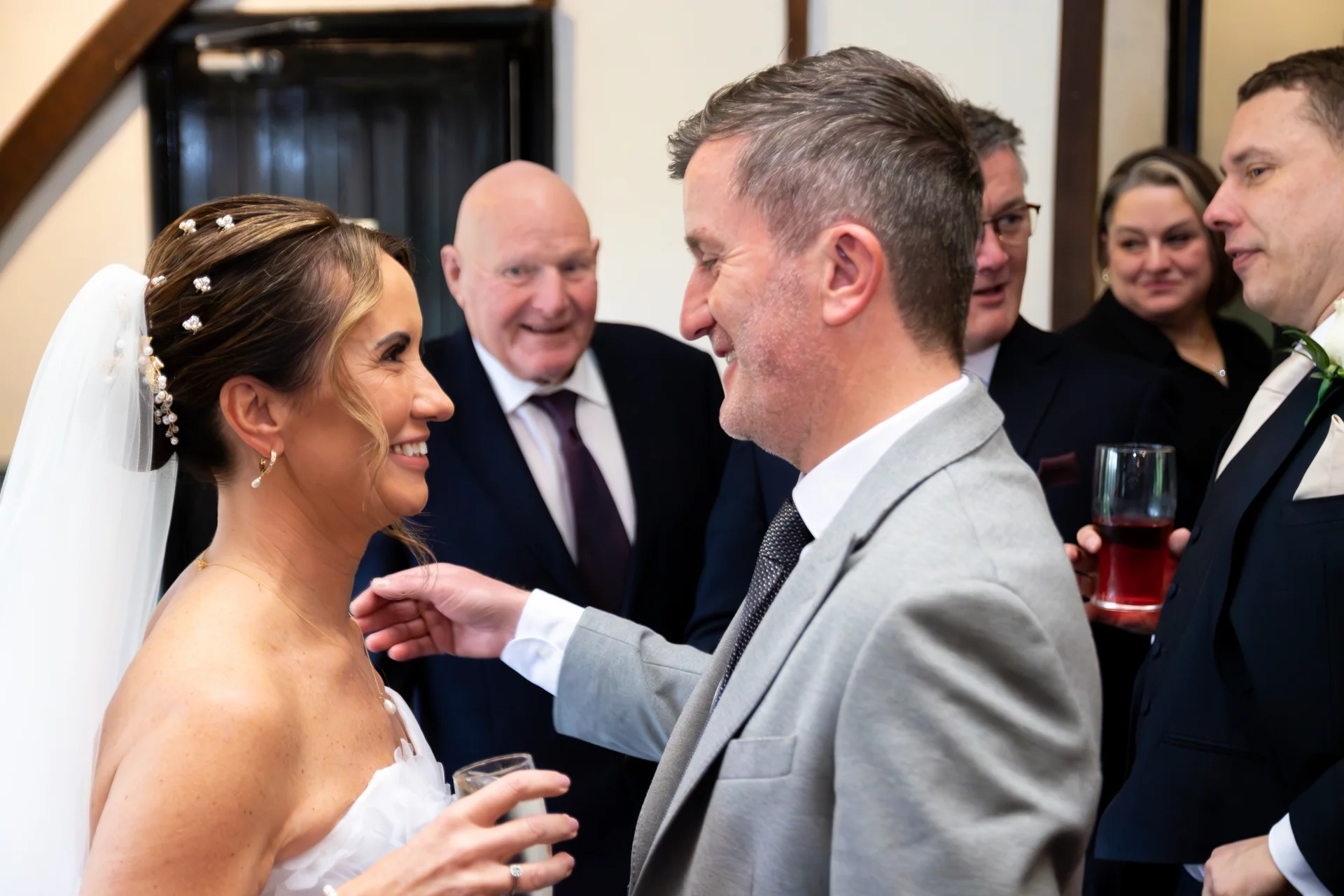A smiling bride and groom share a joyful, tender moment together among mingling wedding guests indoors.