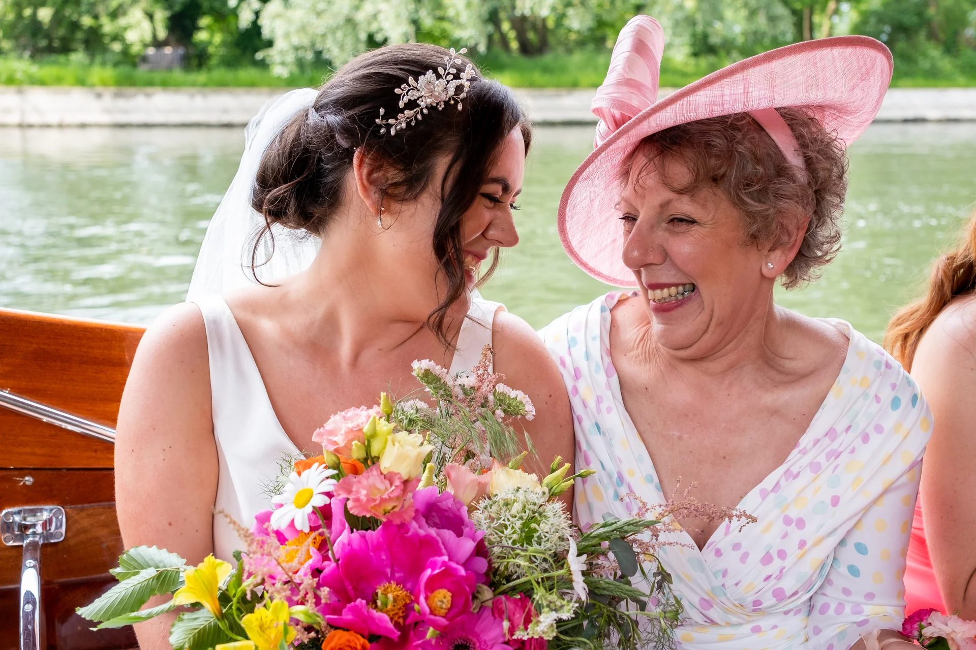 A joyful bride and an older guest wearing a large pink hat share a happy, laughing moment while seated together on a wooden boat holding a colourful floral bouquet.