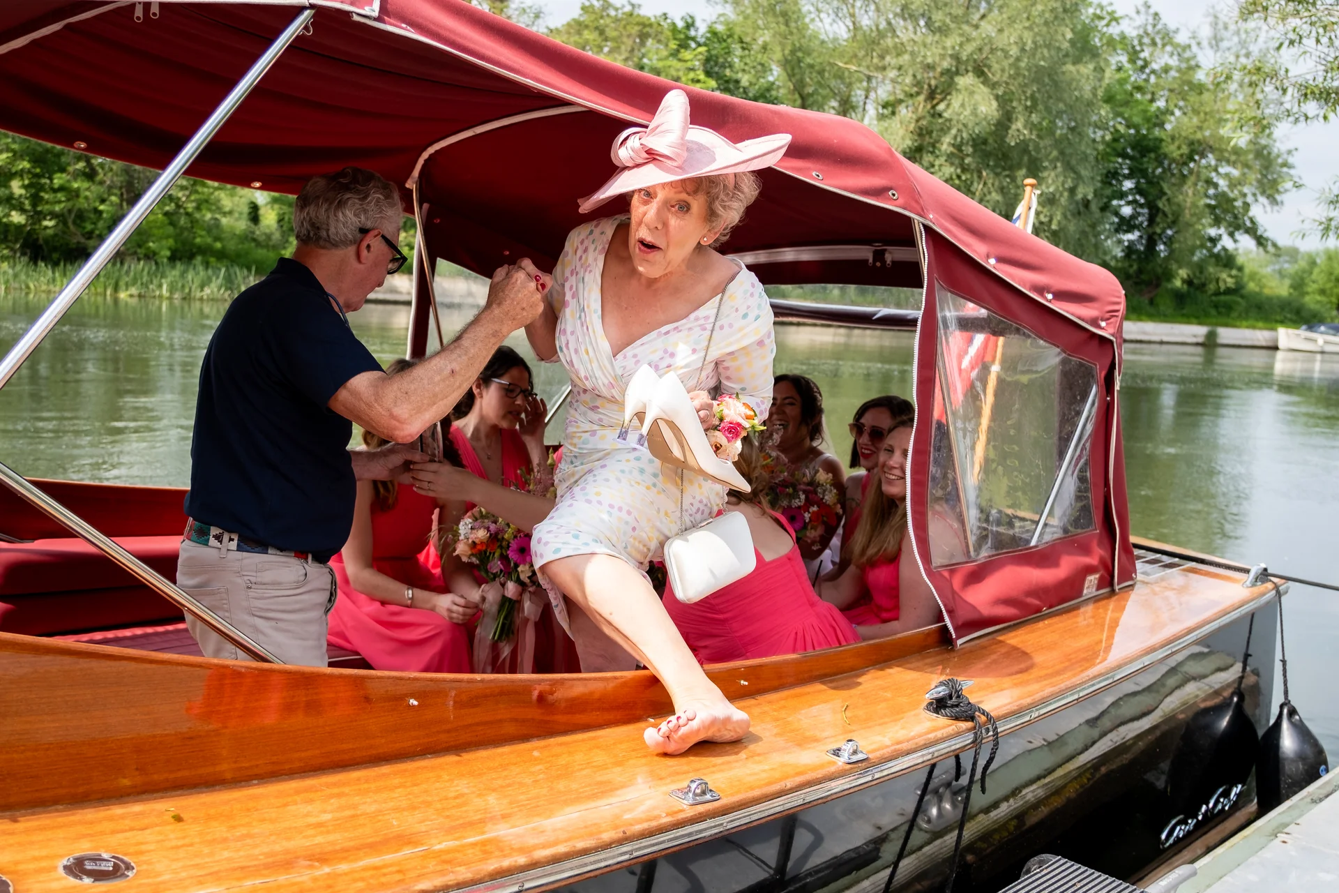 An elderly woman wearing a pink fascinator steps barefoot off a wooden boat while holding her heels, assisted by a man on the dock.