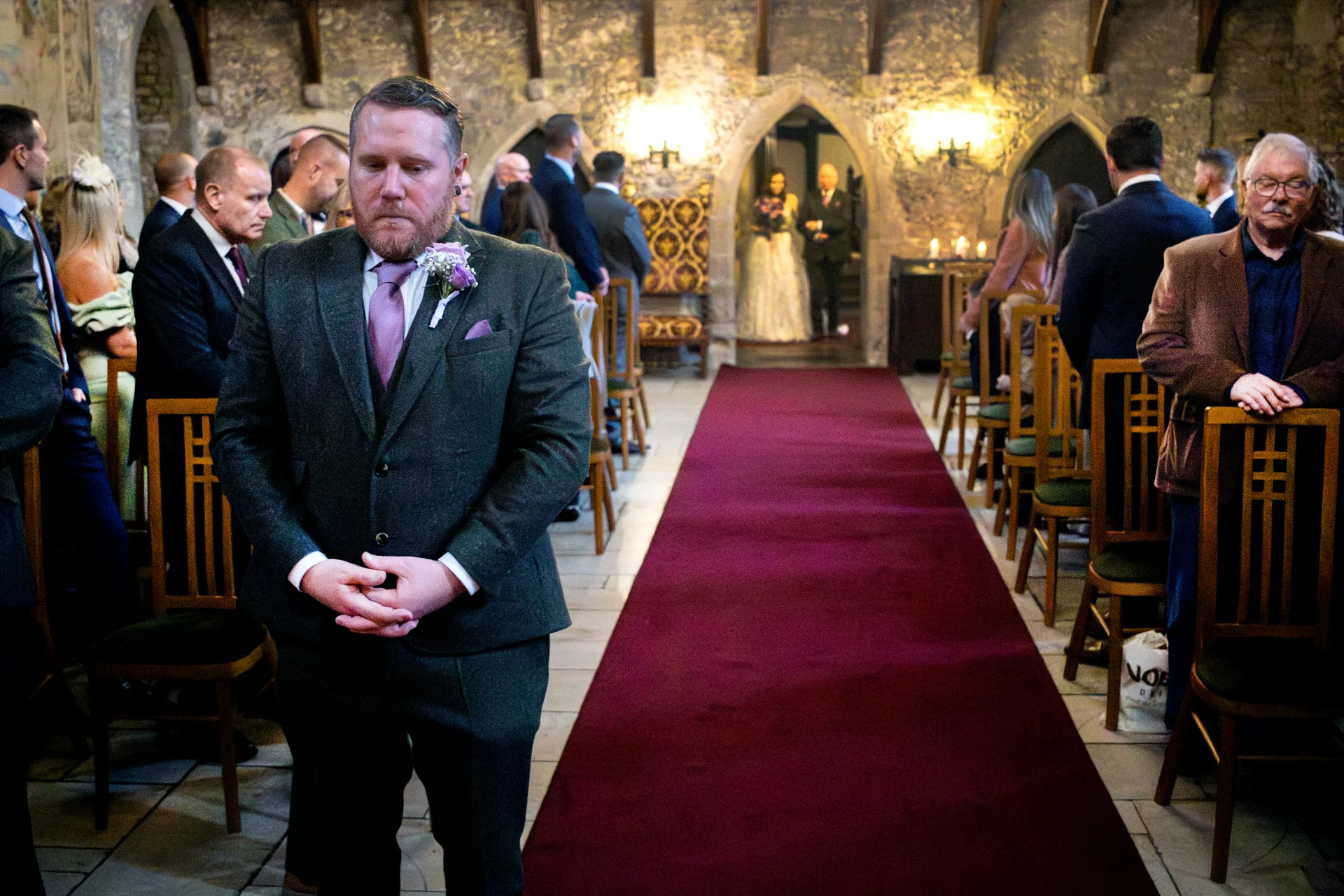 An emotional groom waits patiently at the front of a historic stone castle venue as guests look on and the bride prepares to walk down the red carpeted aisle.