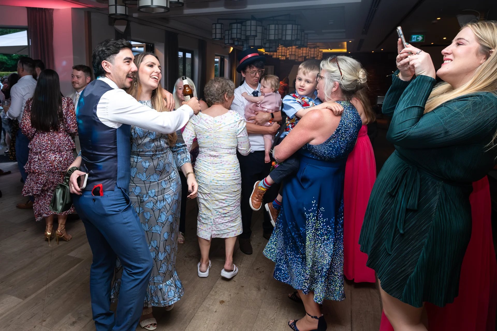 Wedding guests enjoy a lively reception inside a hotel venue, with people laughing, dancing, and capturing memories on their phones.