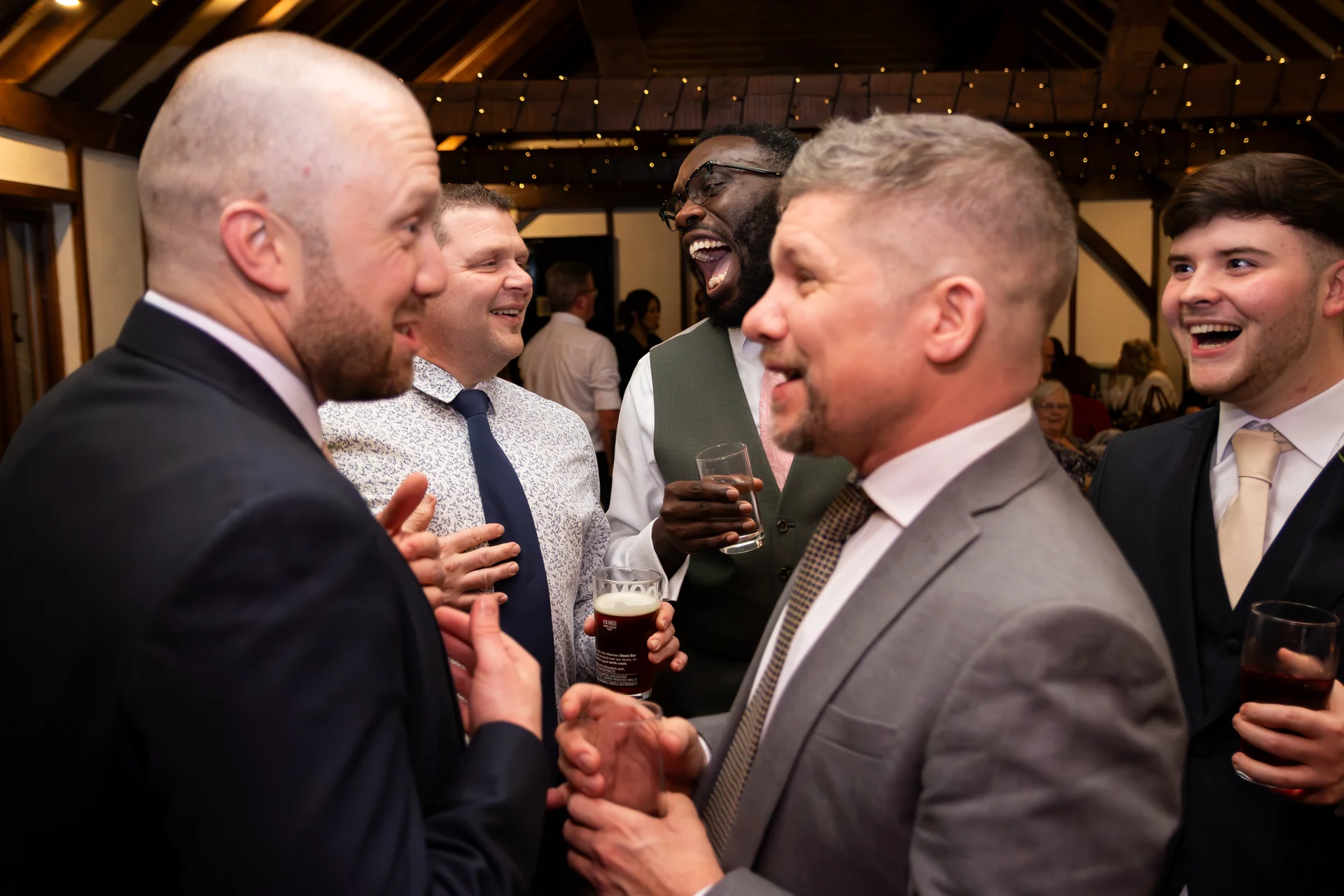 A group of male wedding guests in suits and patterned shirts share a joyful conversation and hearty laugh indoors under rustic exposed rafters.