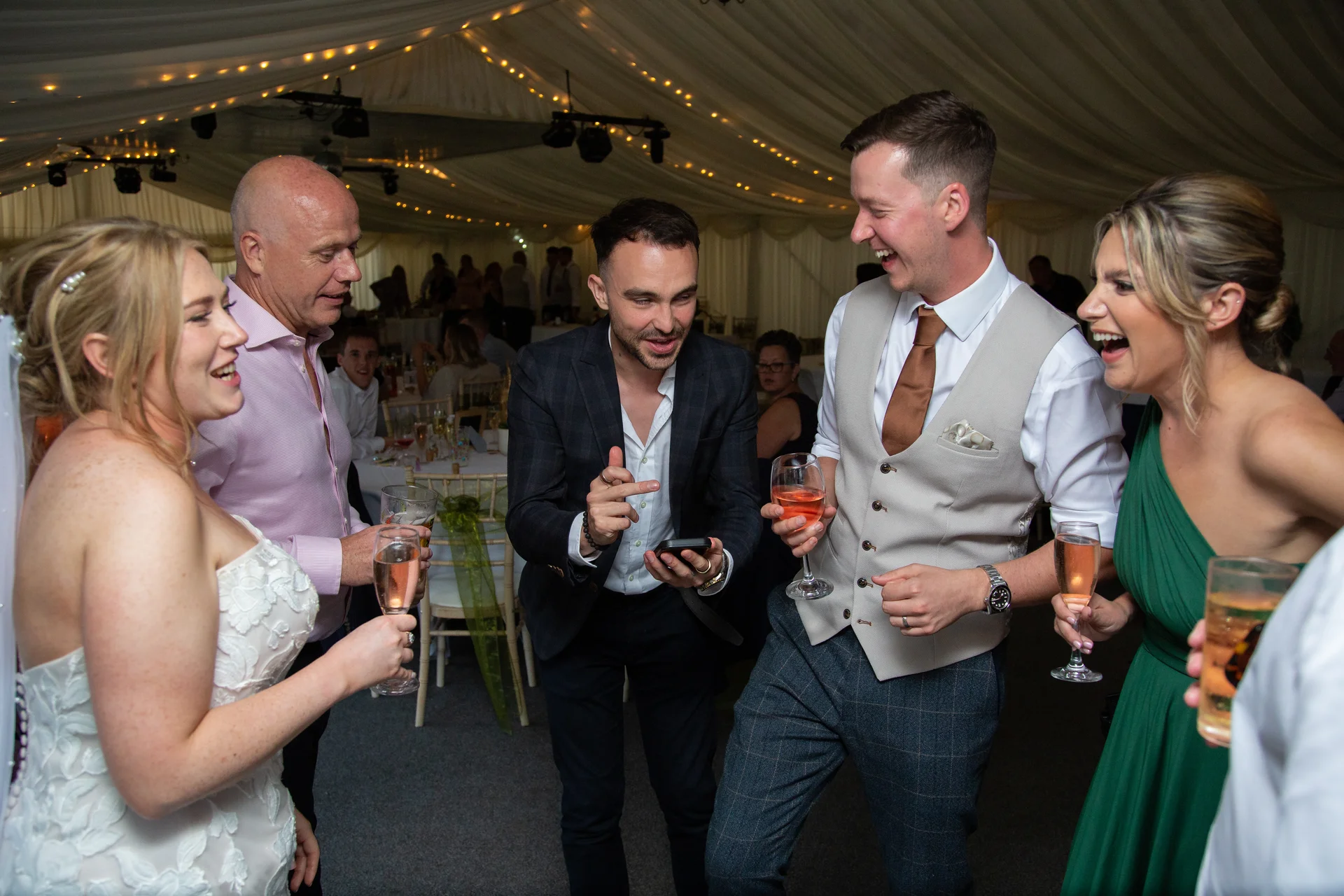 A group of wedding guests laughing and holding drinks inside a marquee lit with fairy lights as a man shows them something on his phone.