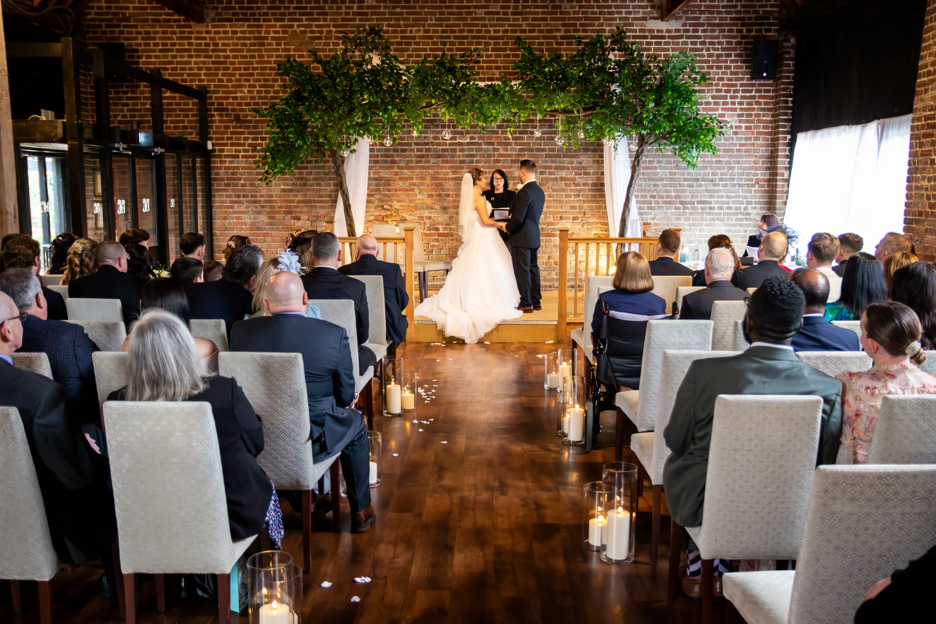 A bride and groom exchange vows at the top of the aisle beneath a decorated tree arch inside a rustic brick barn wedding venue.