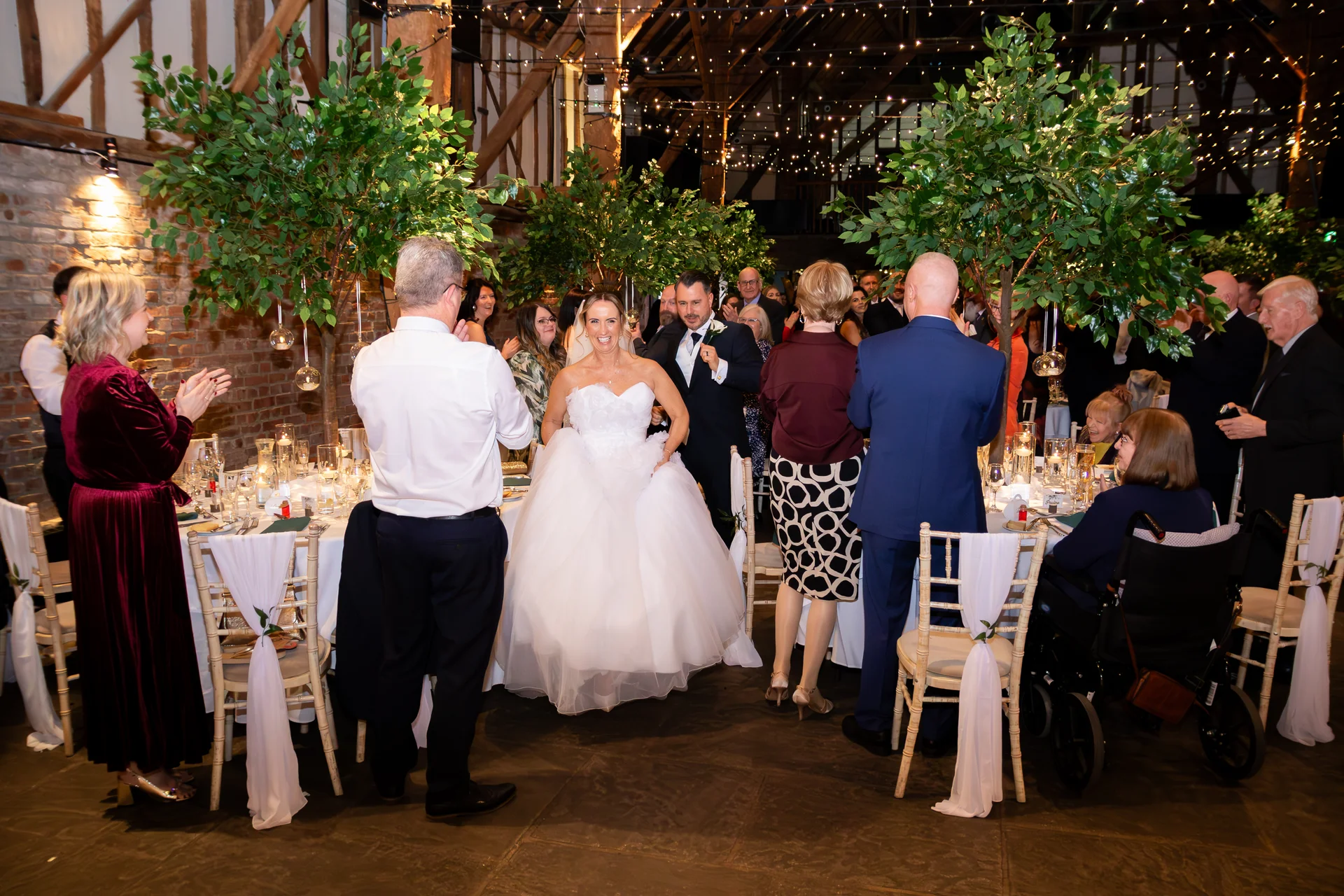 The smiling bride and groom enter a rustic oak-framed barn reception decorated with fairy lights as cheering guests stand to applaud them.