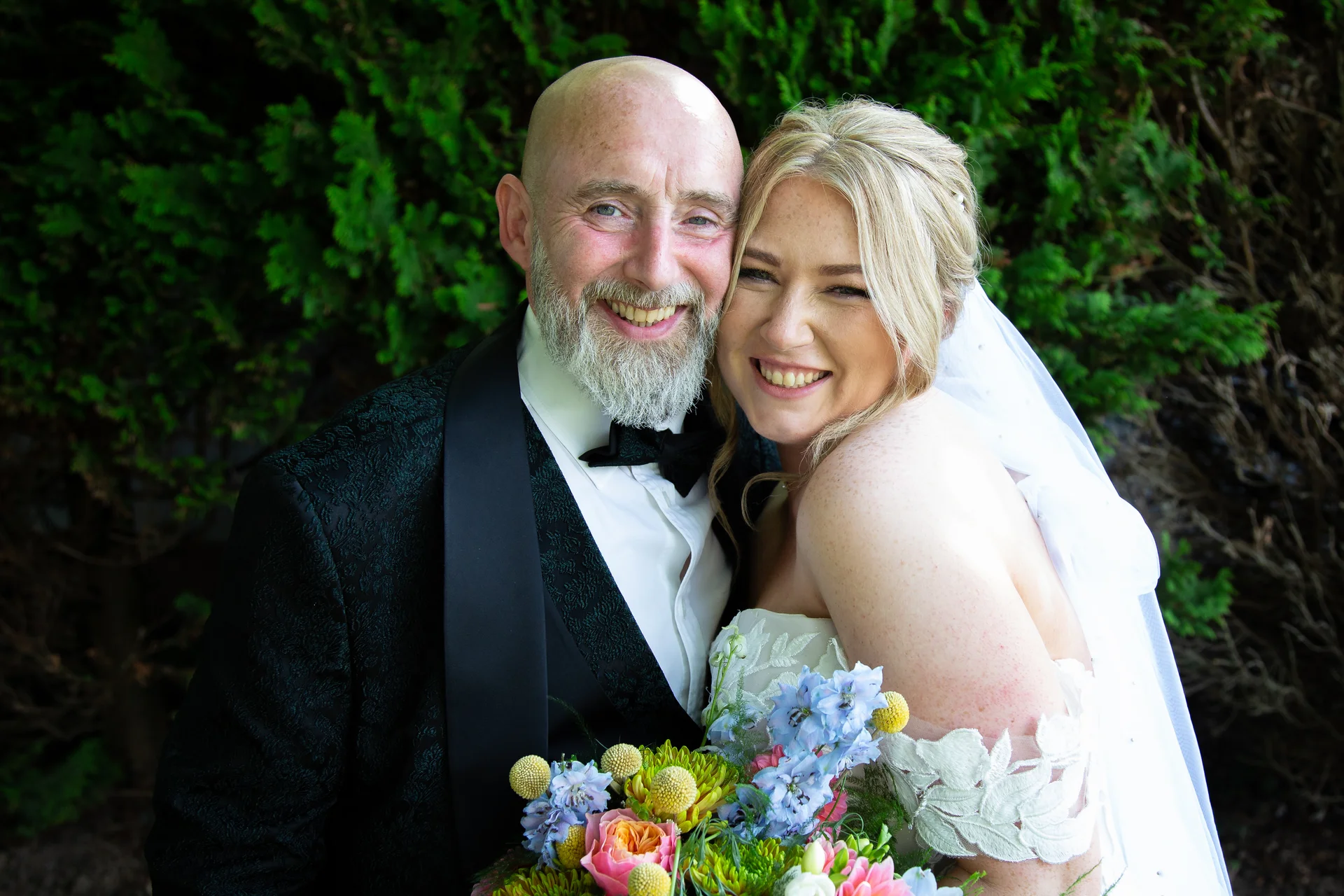 A smiling mature bride and groom pose closely together outdoors in front of lush green foliage, with the bride holding a colourful wildflower bouquet.