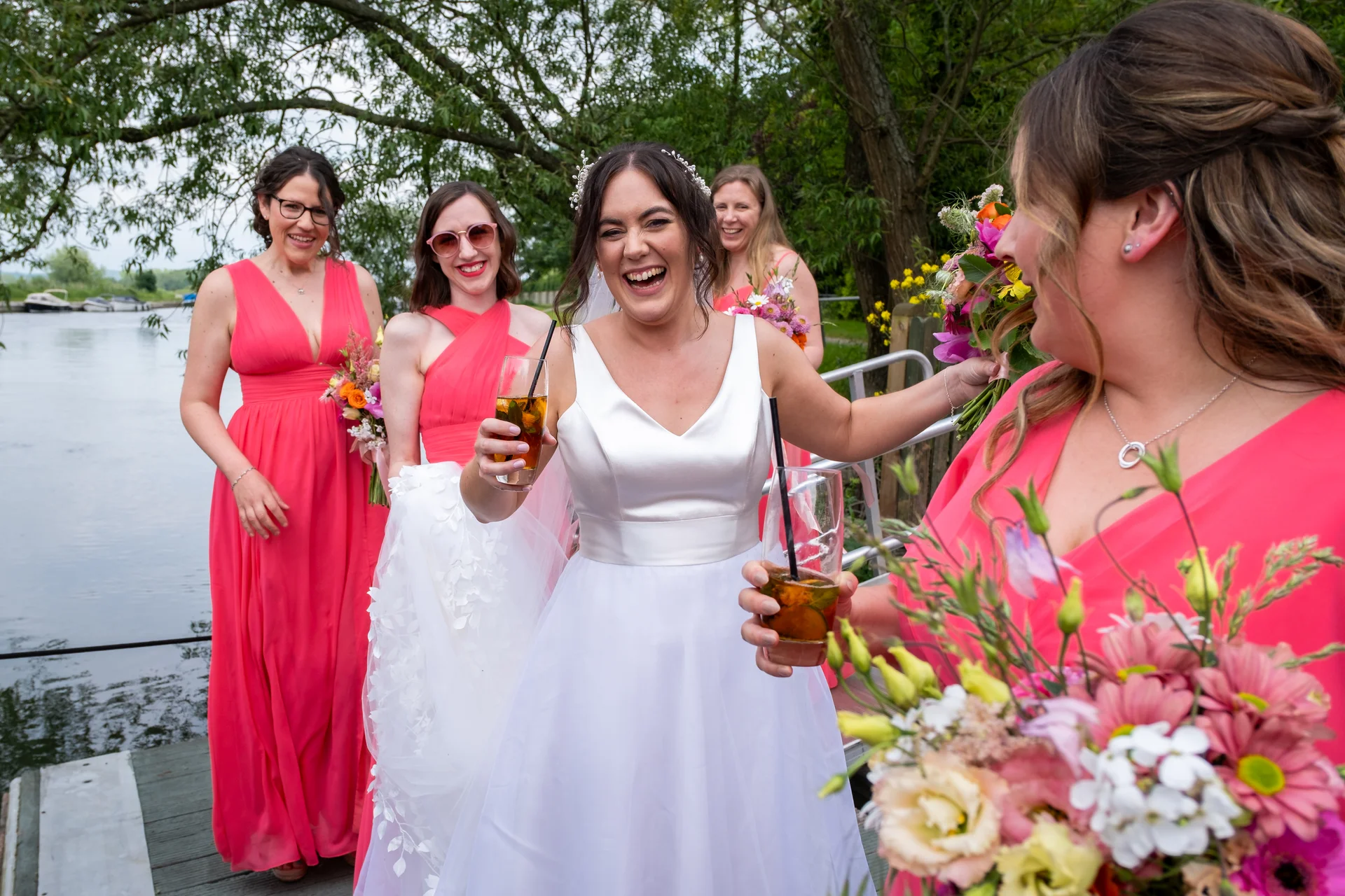 A joyful bride and her bridesmaids wearing pink dresses laugh and hold cocktails by a scenic riverbank.