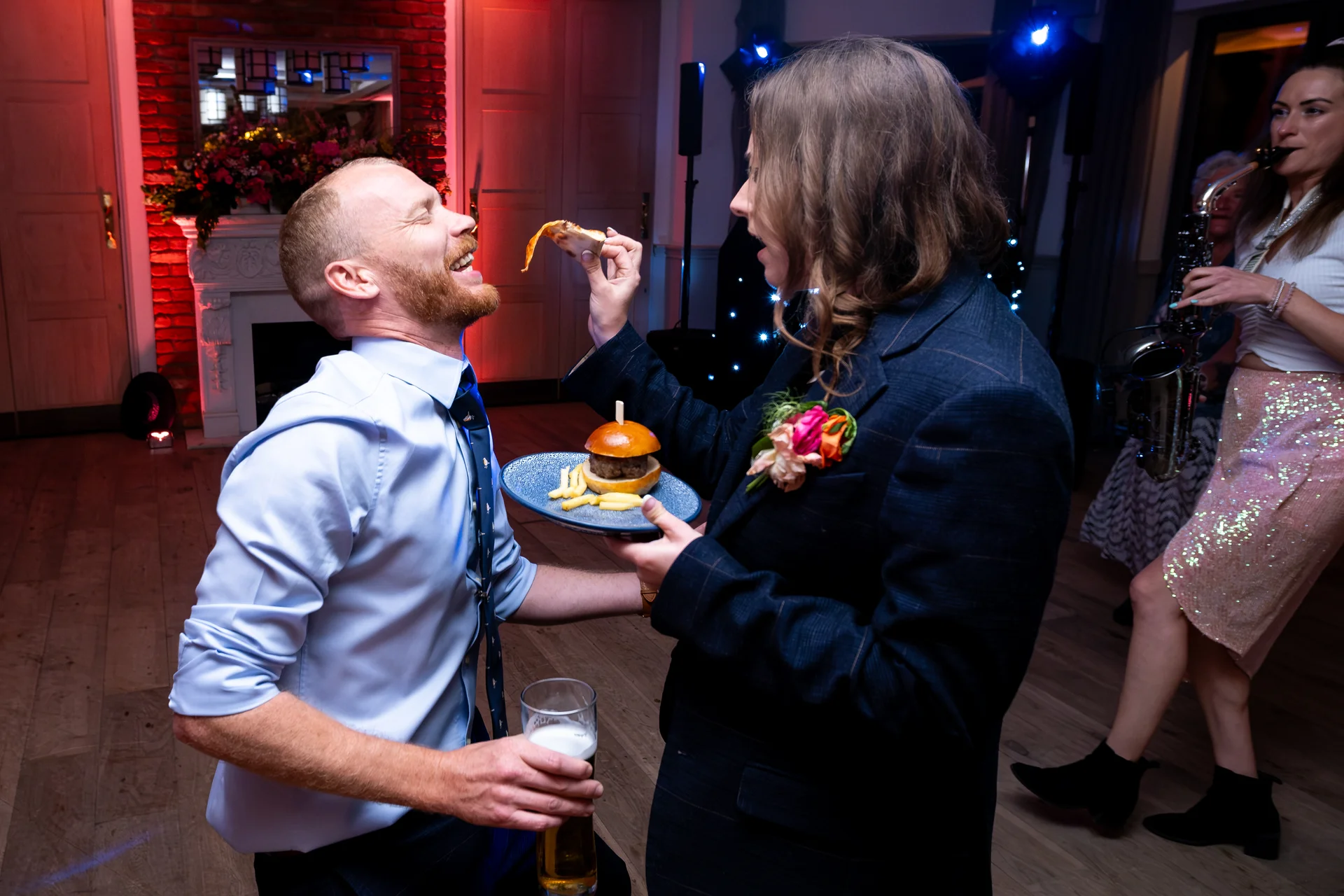 A groom in a blue shirt holds a pint of beer while his partner in a dark patterned suit playfully feeds him a slice of pizza during the evening wedding reception.