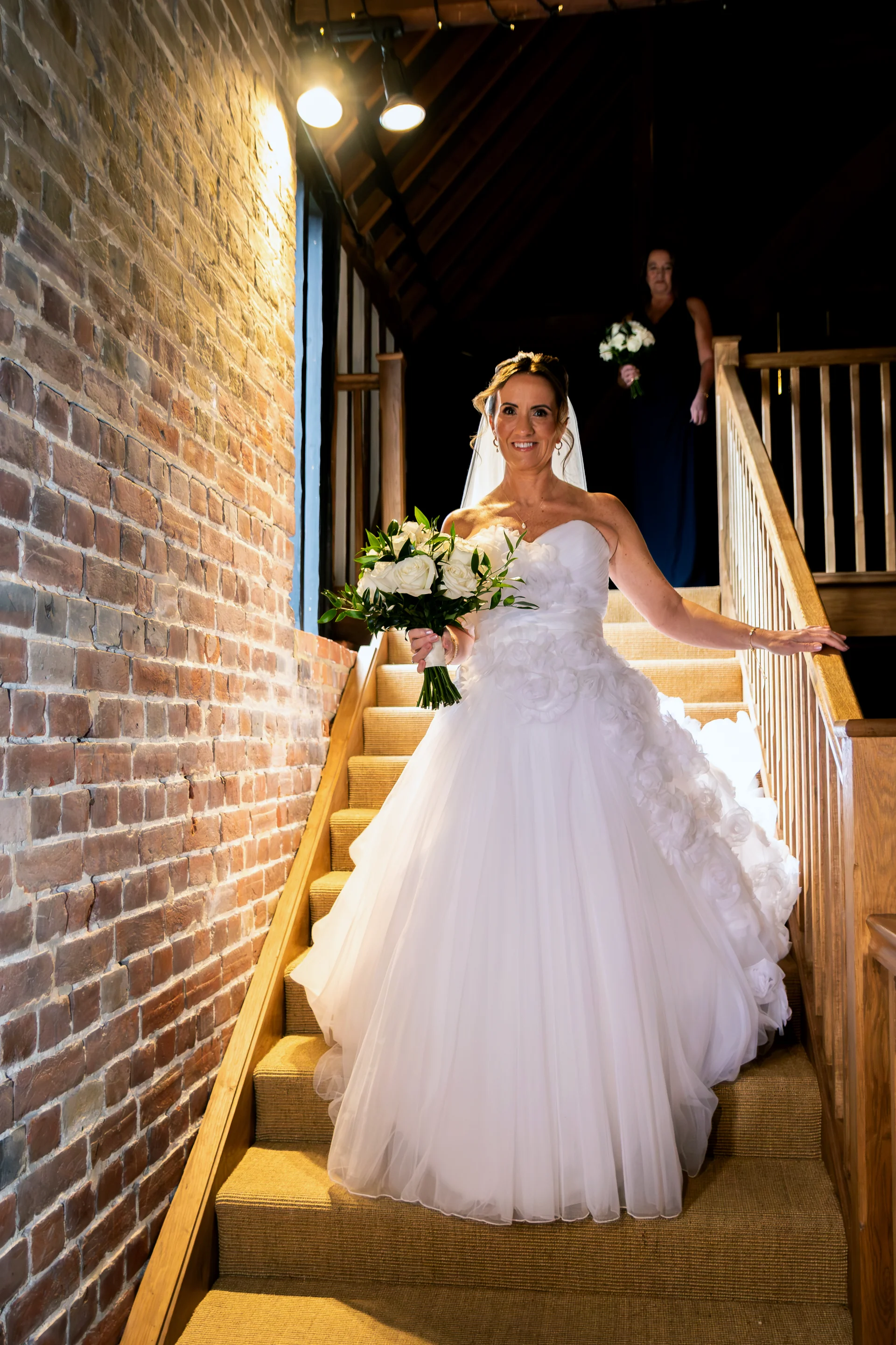 A smiling bride holding a white rose bouquet descends a wooden staircase inside a converted barn with exposed brick walls.