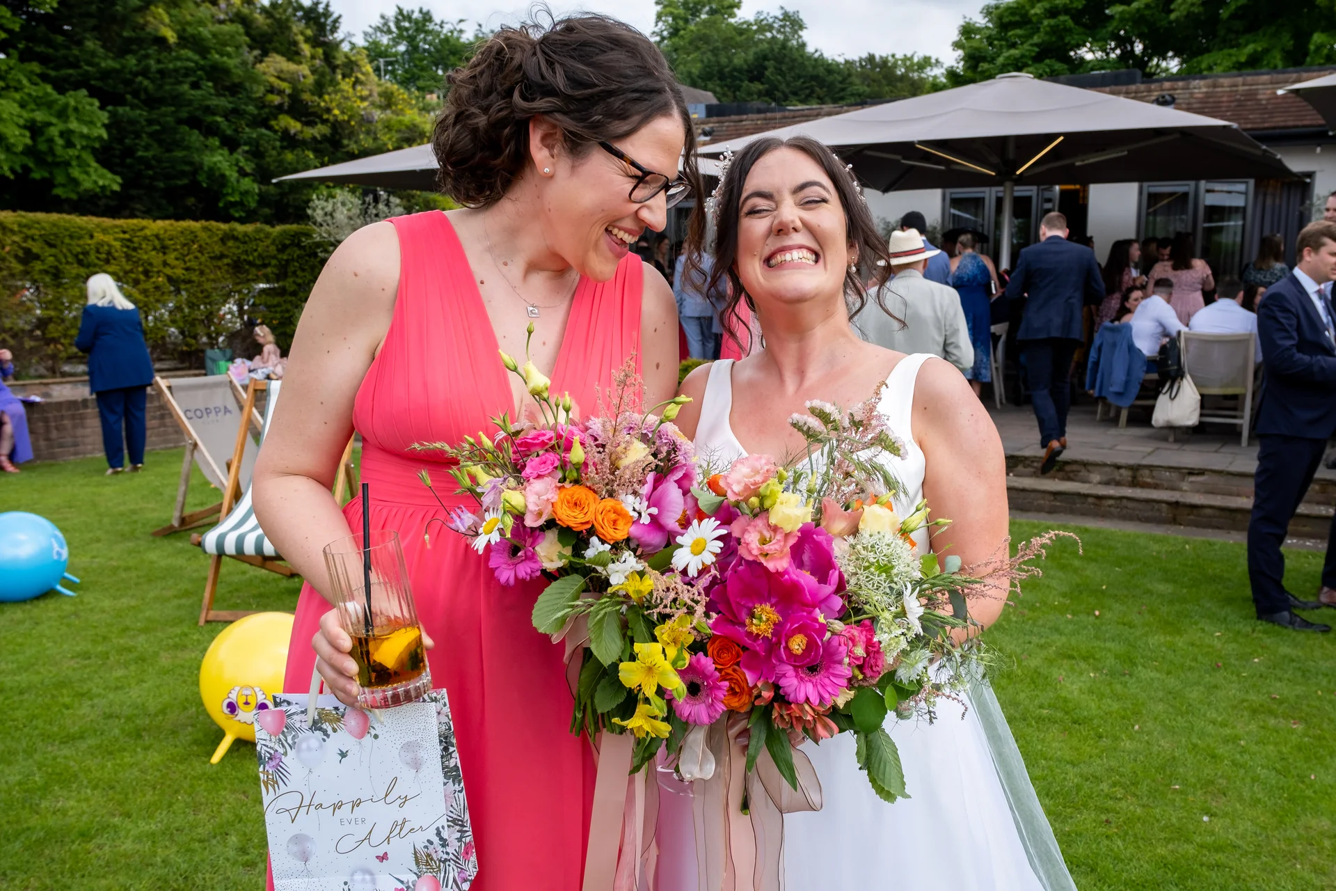 Two smiling brides share a joyful moment in a sunlit garden, holding an abundant, colourful wildflower bouquet with trailing ribbons.