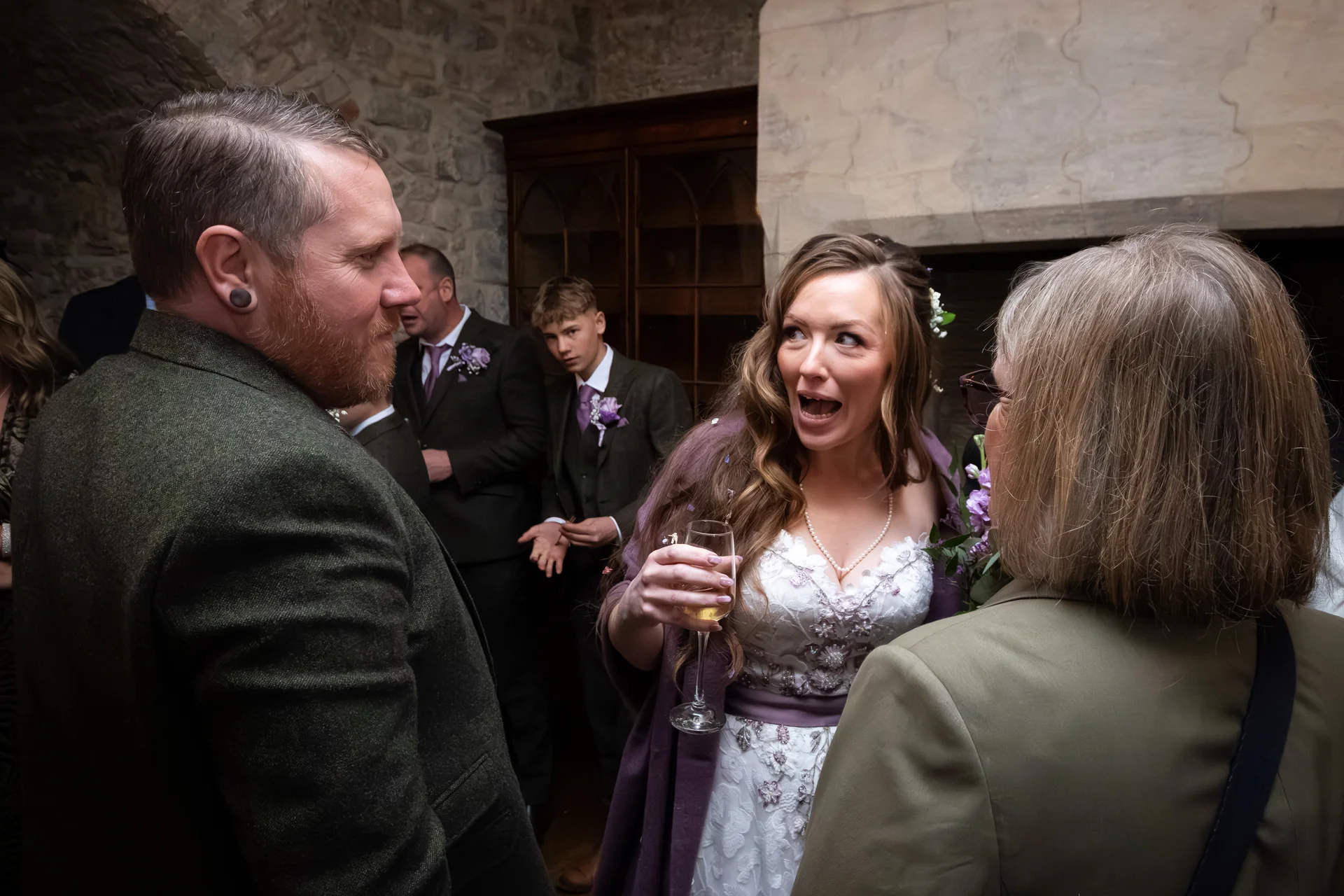 A joyful bride with an expressive expression holds a glass of champagne while chatting with guests in a rustic stone interior during the wedding reception.