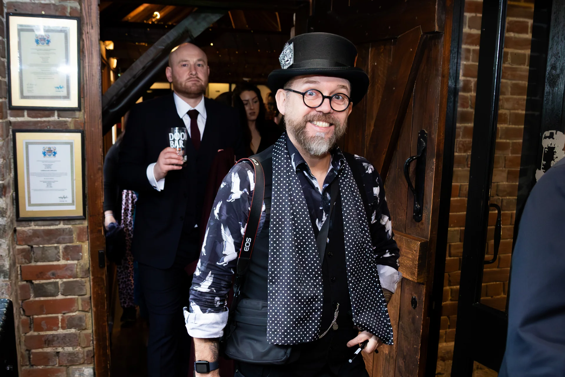 A smiling guest wearing a top hat, patterned shirt, and polka dot scarf walks through a wooden doorway at a barn wedding reception, while other guests follow behind.