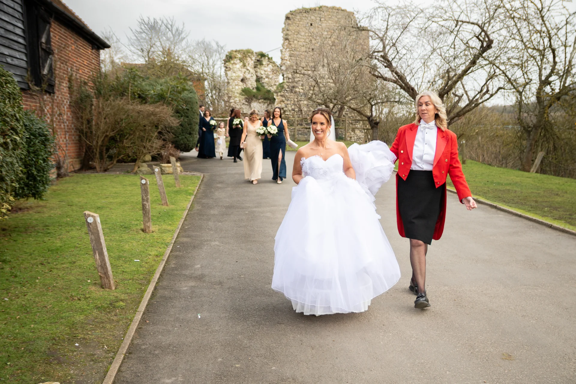A smiling bride holding up her white ball gown walks along an outdoor path alongside a toastmaster in a red jacket, with bridesmaids and castle ruins in the background.
