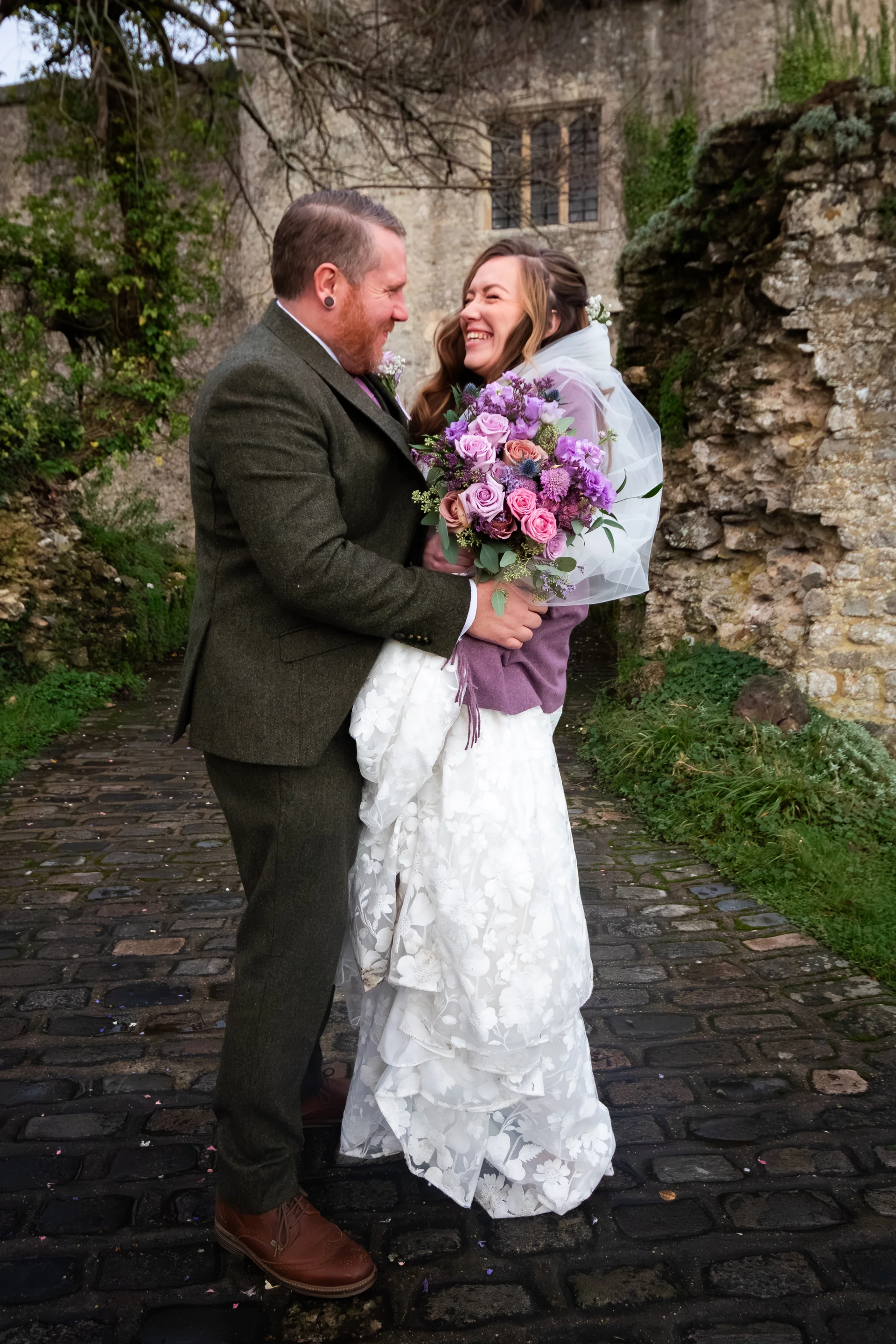 A groom in a green tweed suit joyfully embraces his laughing bride holding a vibrant purple and pink bouquet on a cobbled castle pathway.
