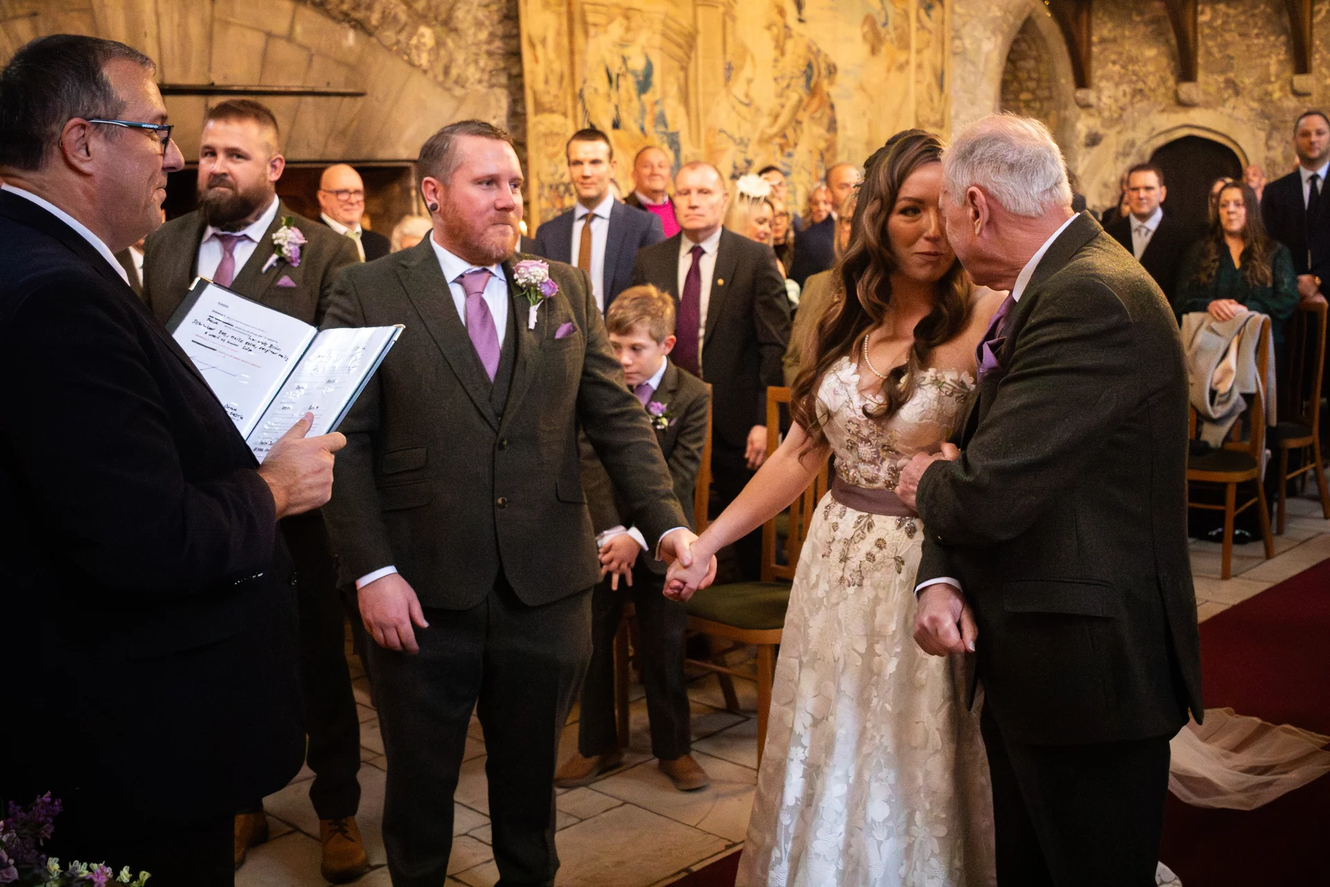 A bride holding hands with her father during a castle wedding ceremony while the groom looks on.