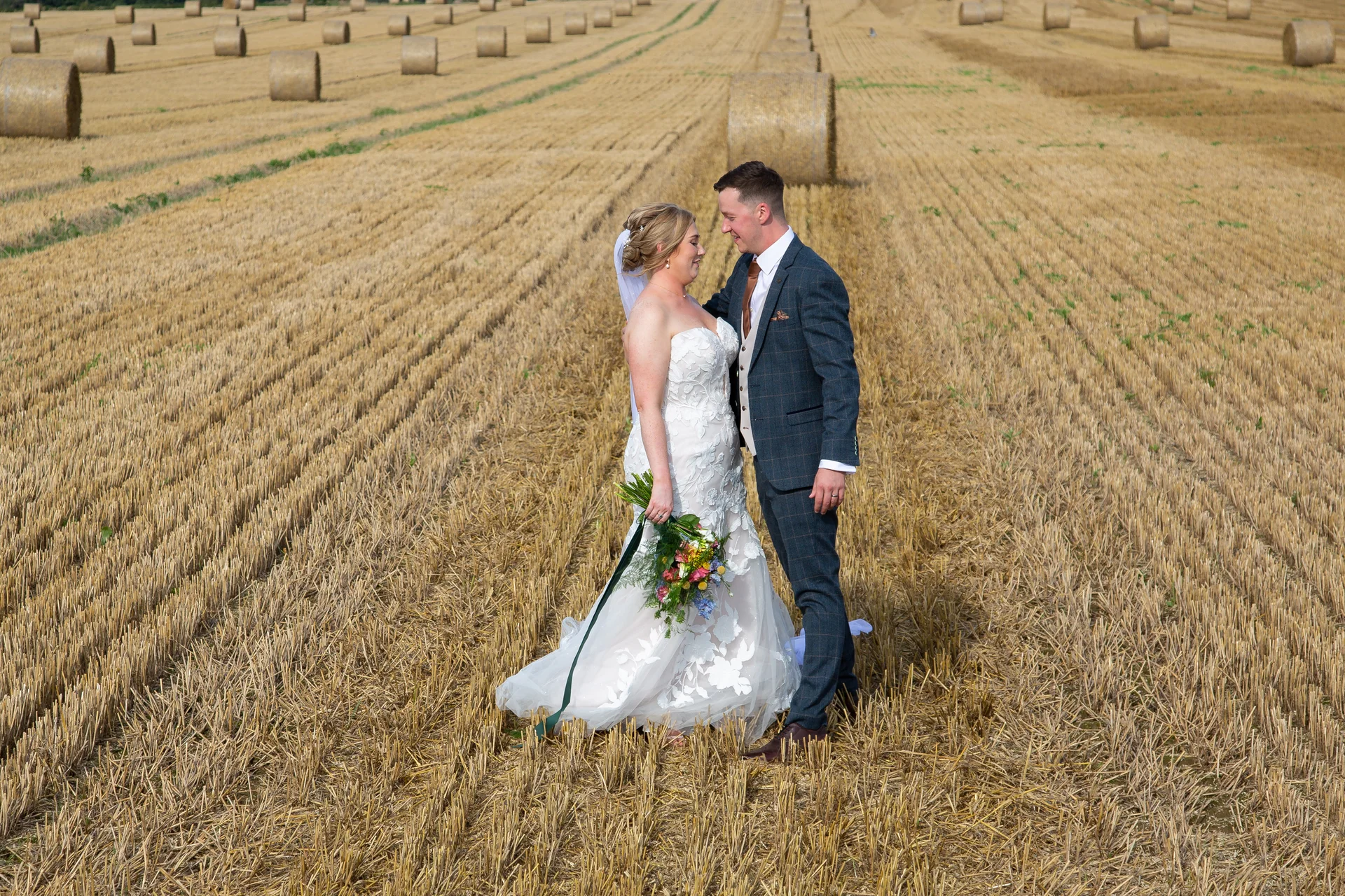 A newlywed couple stands in a harvested golden stubble field scattered with round hay bales, captured from a high-angle aerial view.