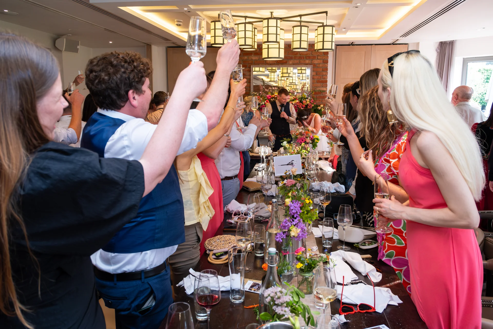 Wedding guests raise their glasses in a toast during speeches at a lively indoor reception with a long banquet table.
