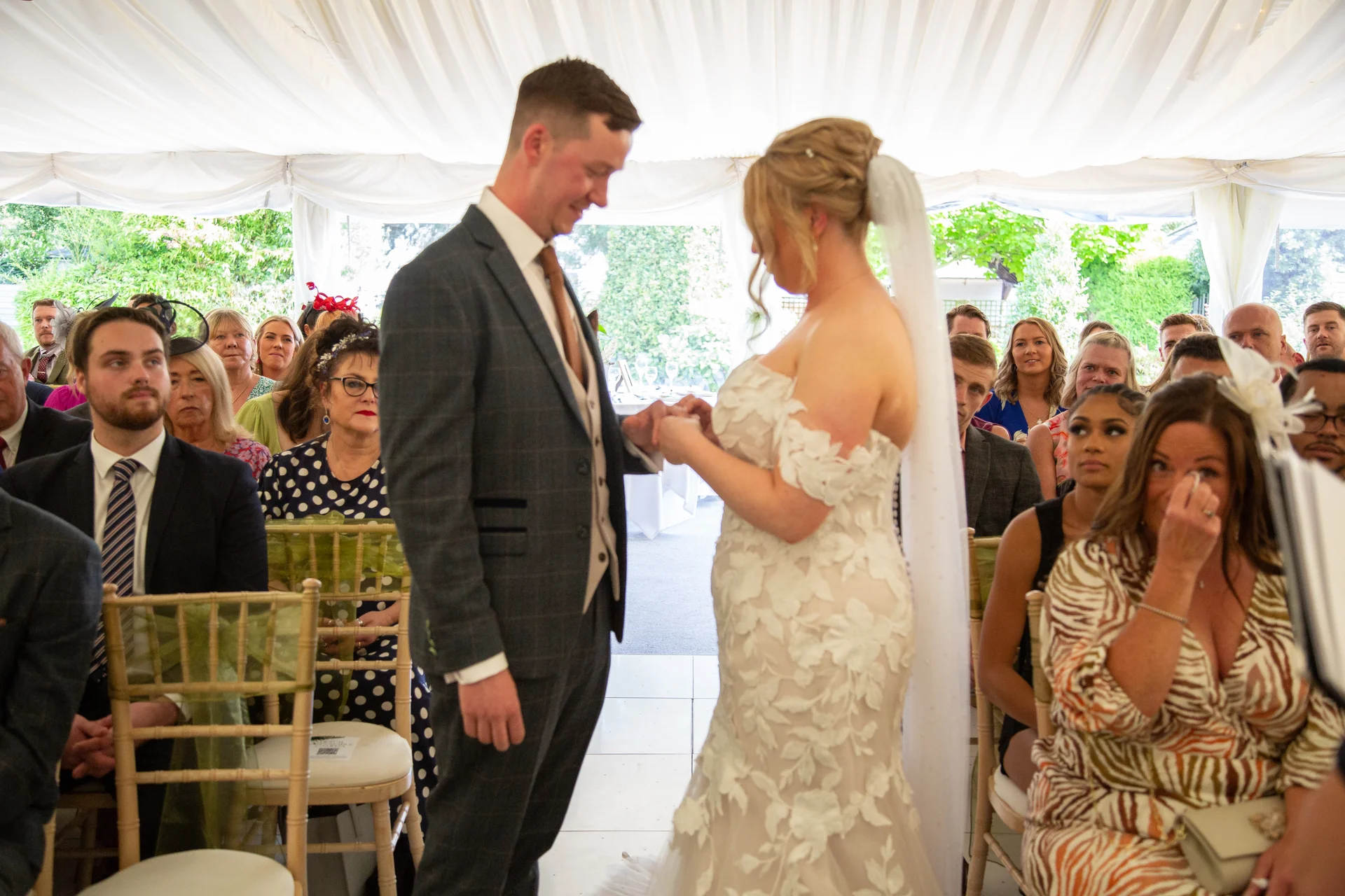 A groom and bride exchange rings during a marquee ceremony while seated guests look on, with one emotional guest wiping away tears in the foreground.