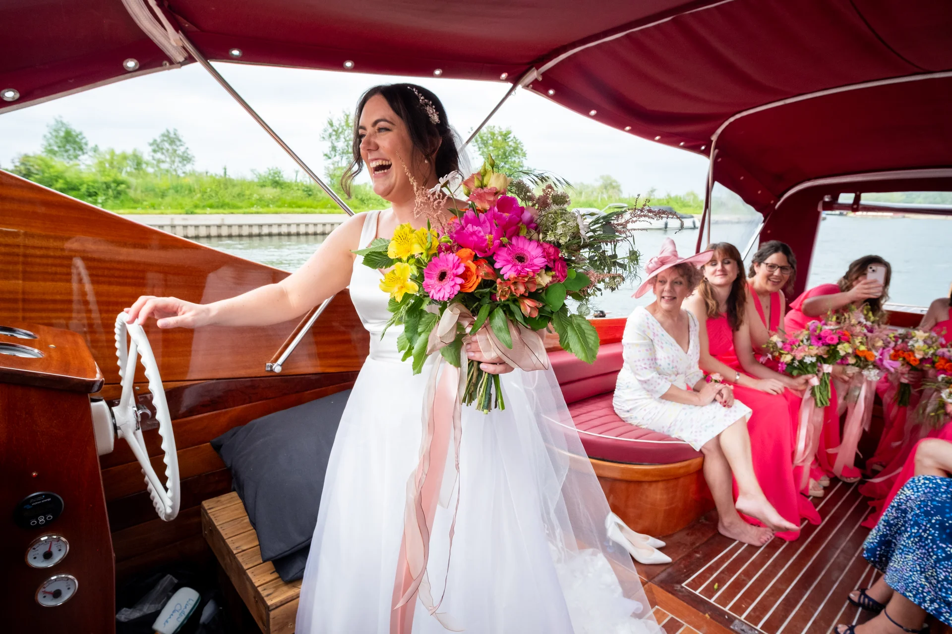 A joyful bride steering a wooden boat while holding a vibrant, colourful bouquet, with bridesmaids and guests laughing in the background.