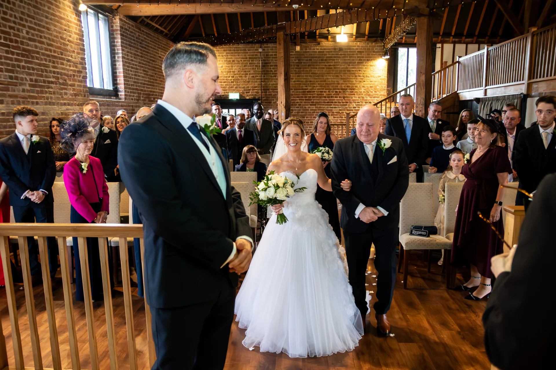 A smiling bride walks down the aisle on her father's arm inside a rustic converted barn with exposed brick walls and wooden beams, towards the waiting groom.
