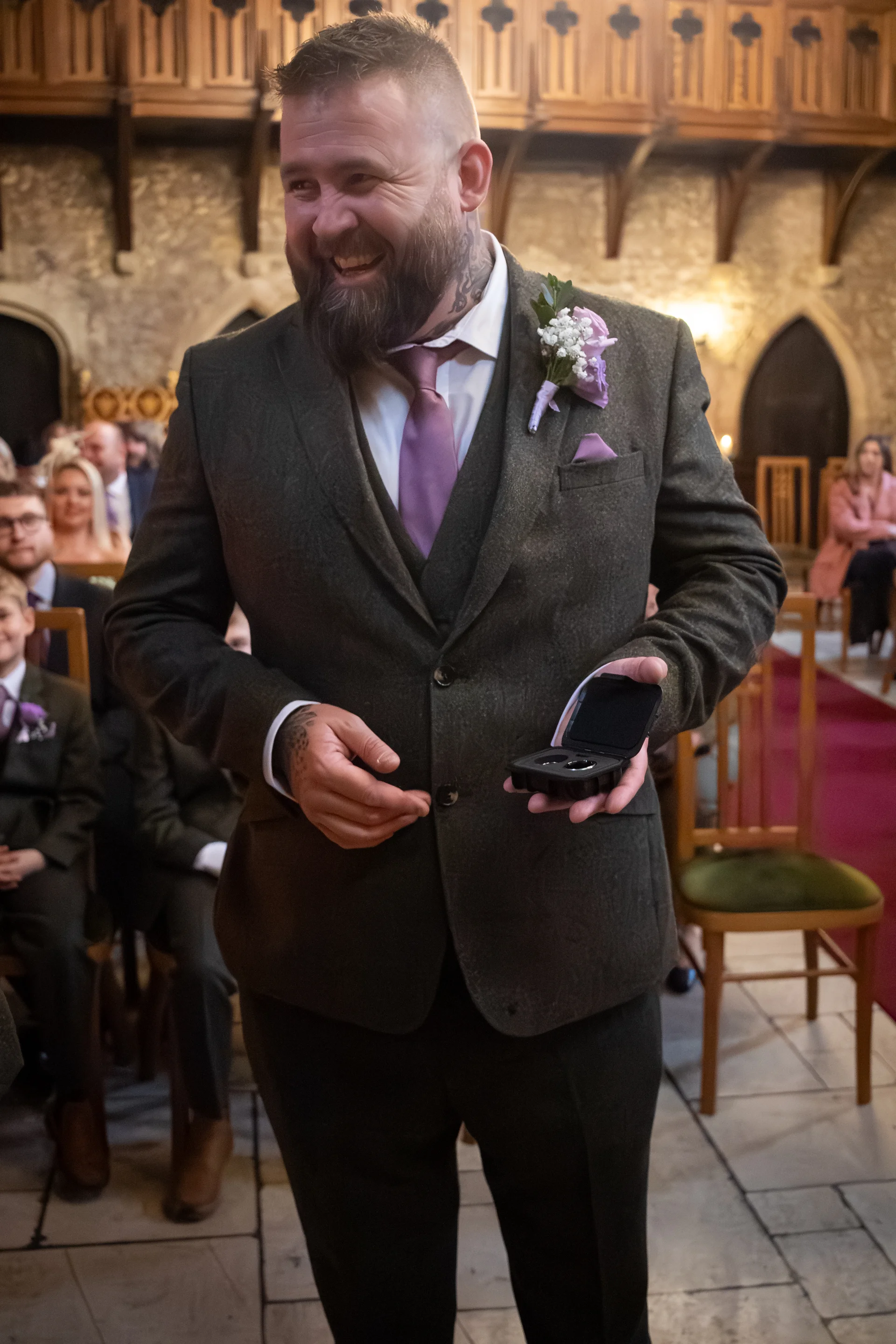 A smiling groom wearing a tweed suit and purple tie holds an open ring box during a wedding ceremony inside a stone church.