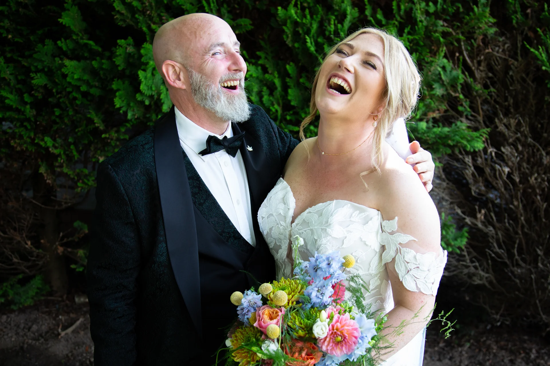An older couple in wedding attire laugh joyfully together outdoors in front of green foliage, holding a vibrant colourful bouquet.