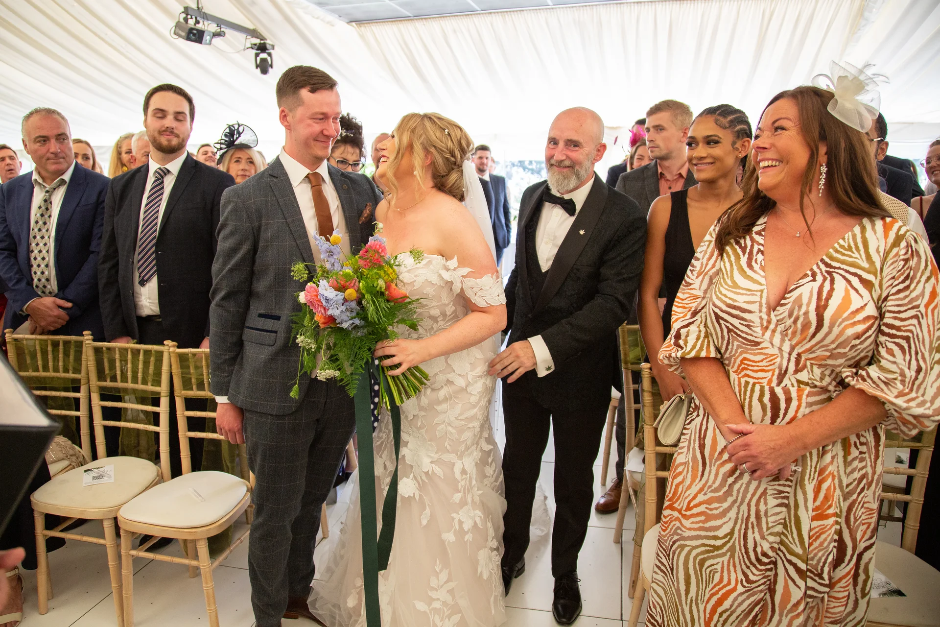 The bride and groom exchange a joyful glance holding a colourful hand-tied bouquet during their marquee ceremony surrounded by laughing guests.