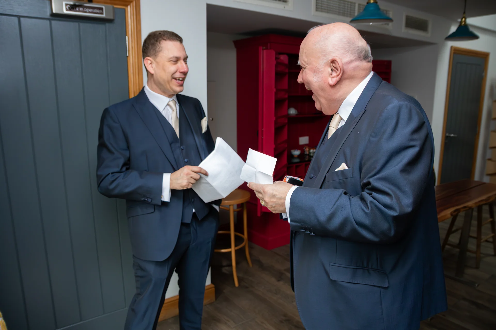 A smiling groom and an older gentleman in matching navy three piece suits laugh heartily while holding notes indoors before the wedding ceremony.