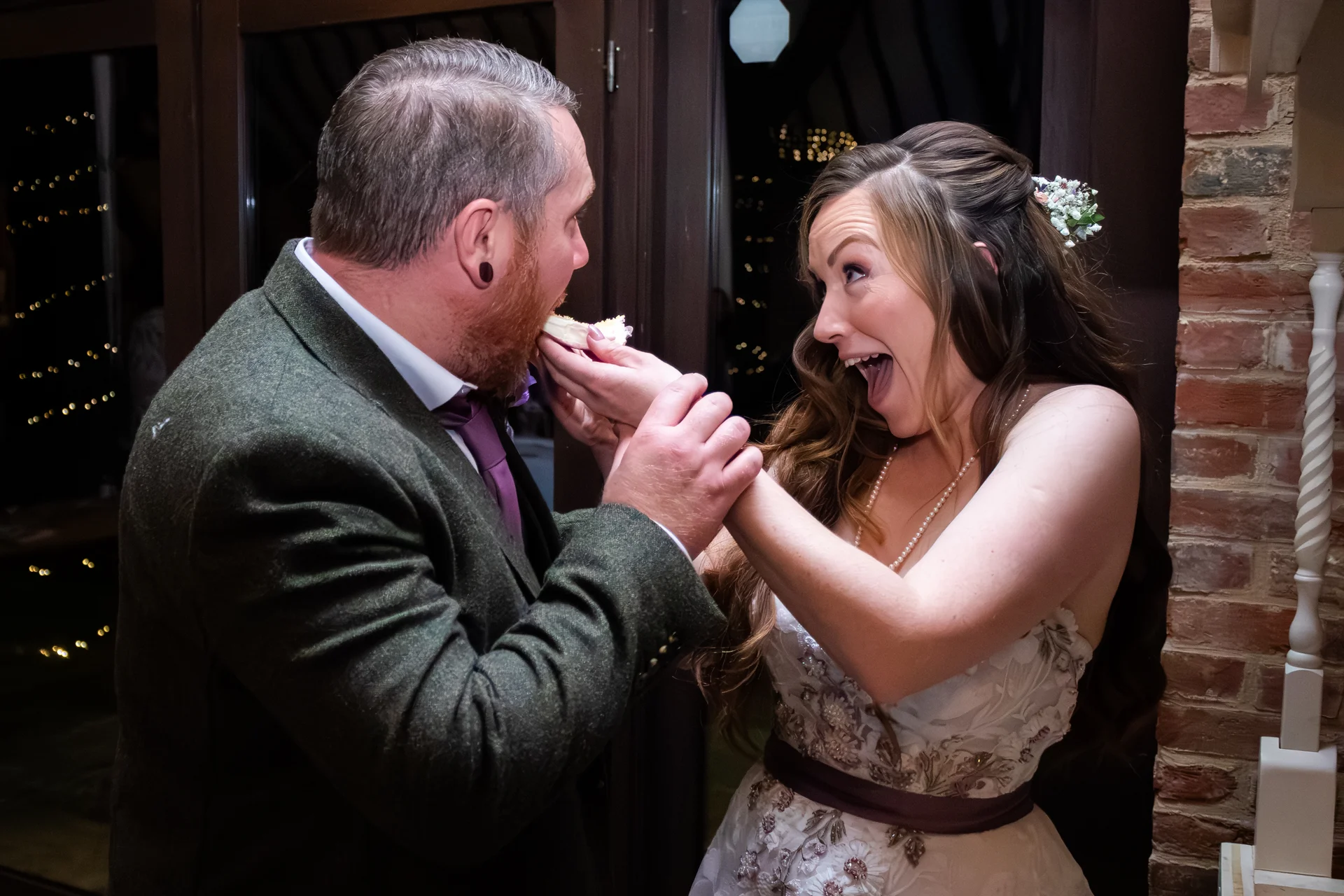 A groom playfully feeds cake to an ecstatic bride wearing a floral applique gown during their wedding evening reception.