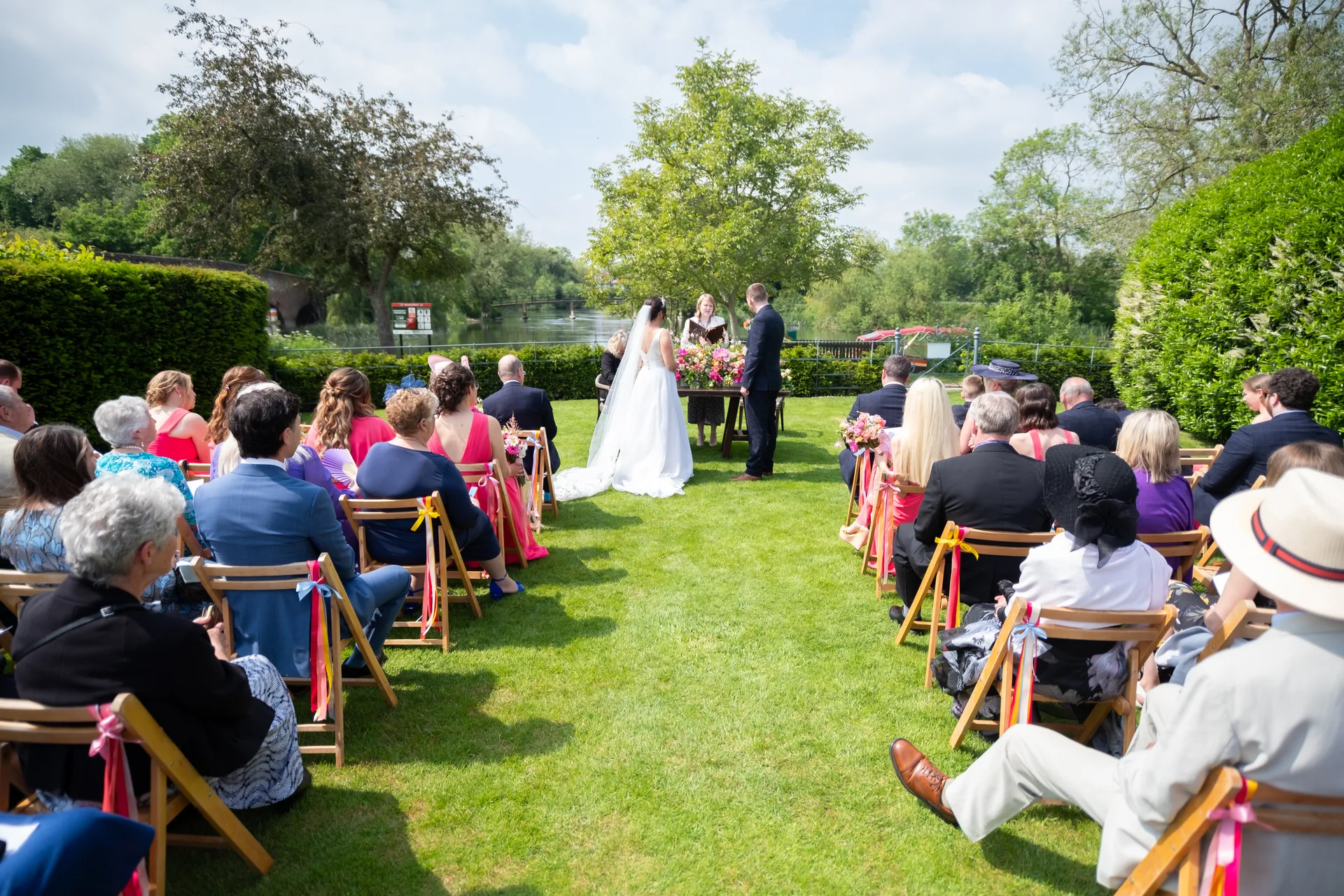 An outdoor wedding ceremony takes place on a sunlit lawn by a river, with the bride and groom standing before a celebrant surrounded by seated guests.