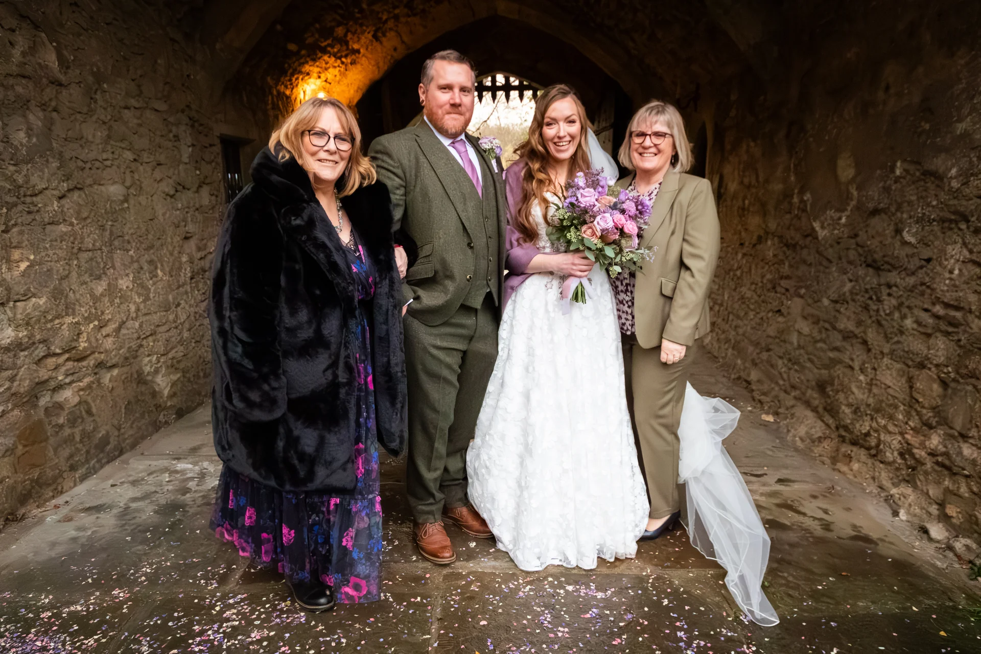 A newlywed couple poses inside a historic stone castle archway alongside family members, surrounded by confetti scattered on the ground.