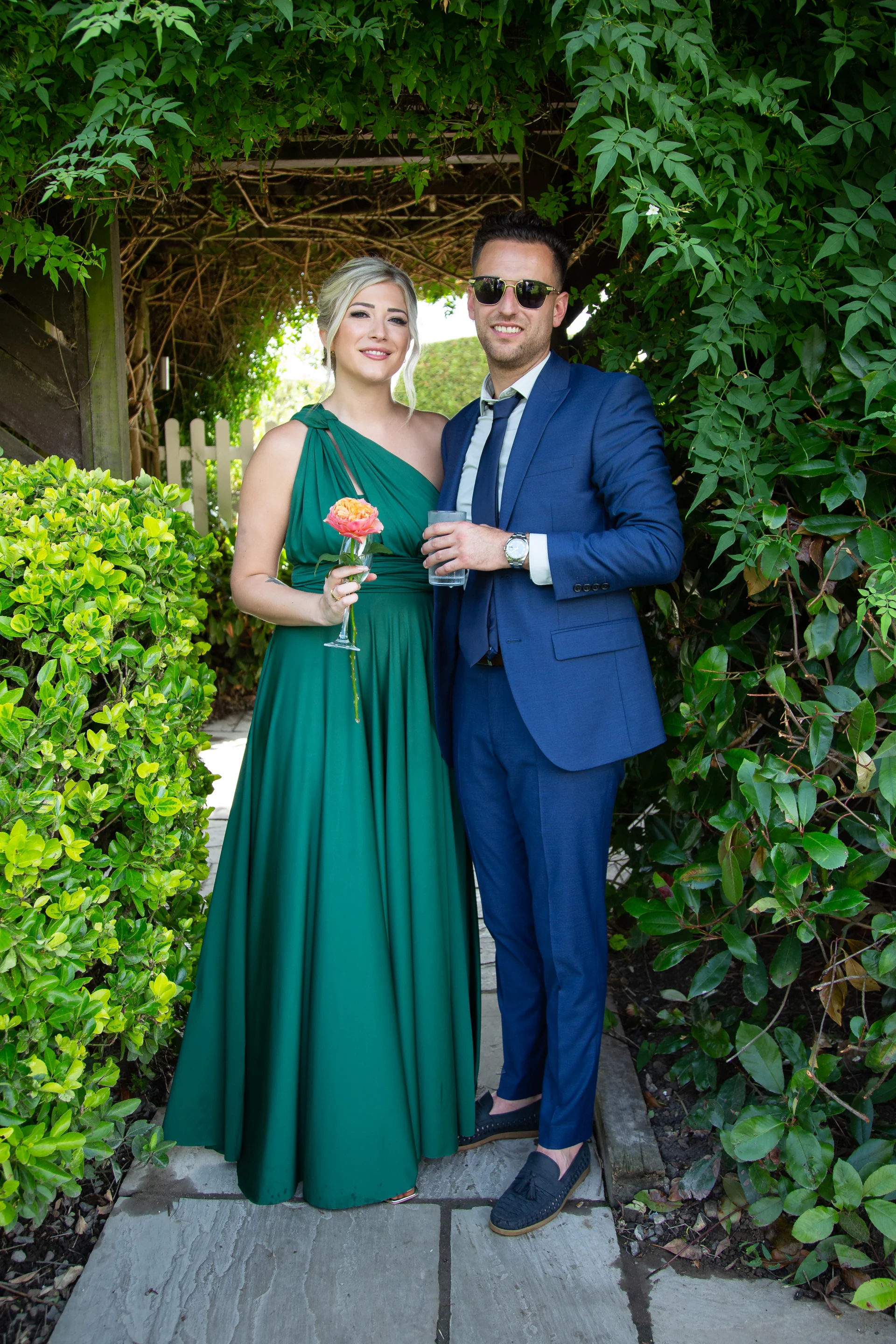 A stylish couple posing together in a lush garden setting, with the bride wearing a striking emerald green one-shoulder gown holding a single rose and the groom in a sharp blue suit with sunglasses.