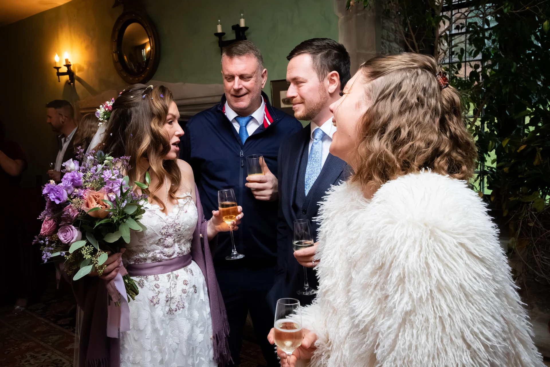 A smiling bride holding a purple floral bouquet chats with wedding guests holding glasses of champagne during the reception drinks.