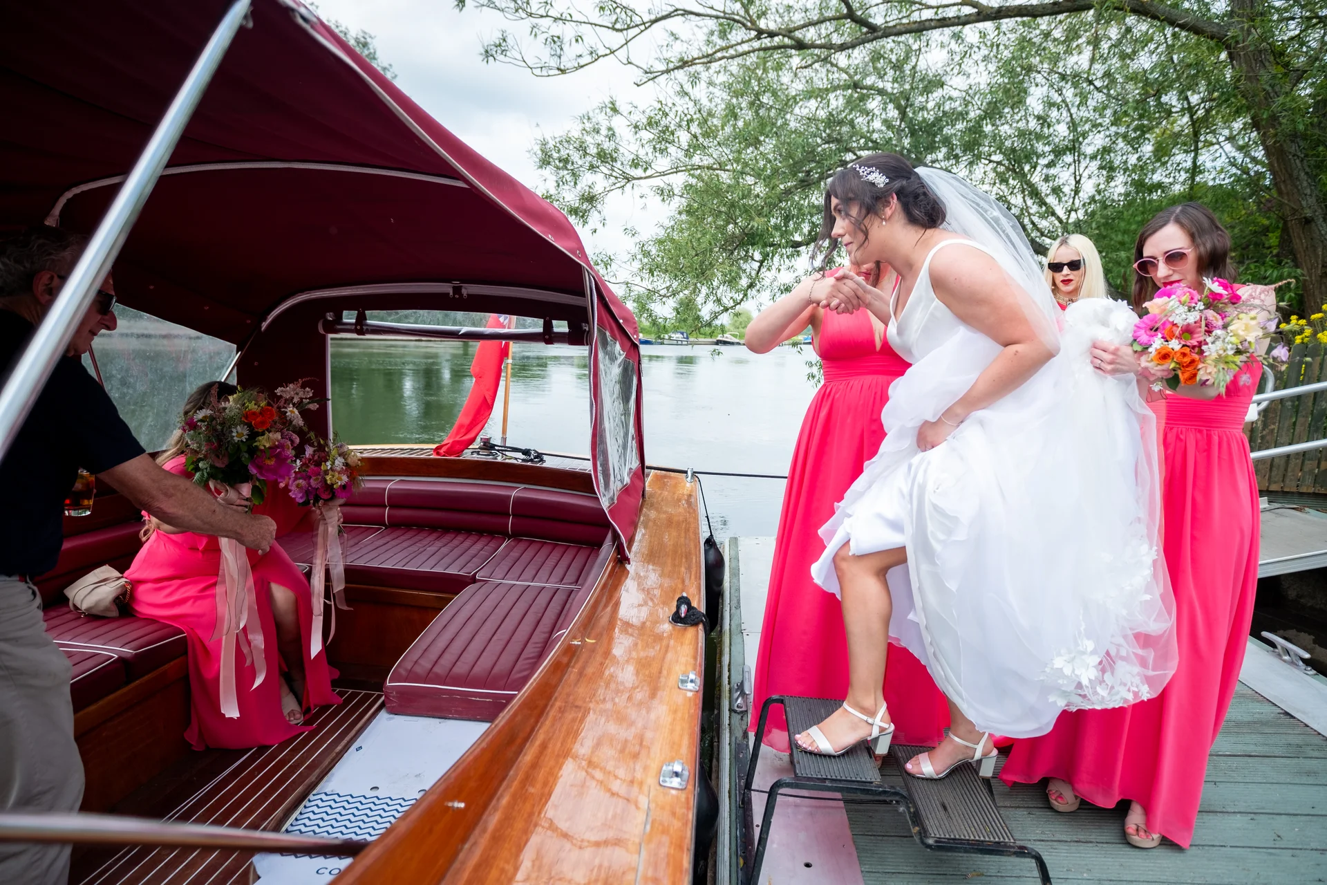 A bride in a white gown and veil steps off a wooden boat onto a jetty, assisted by her bridesmaids in bright pink dresses.