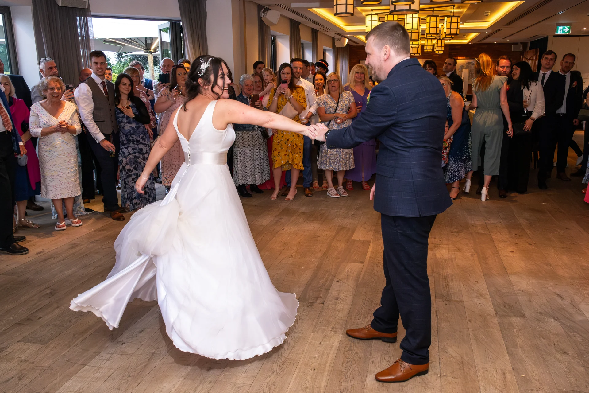 Newlyweds share their joyful first dance in a spacious hotel ballroom while wedding guests look on and capture the moment on their phones.