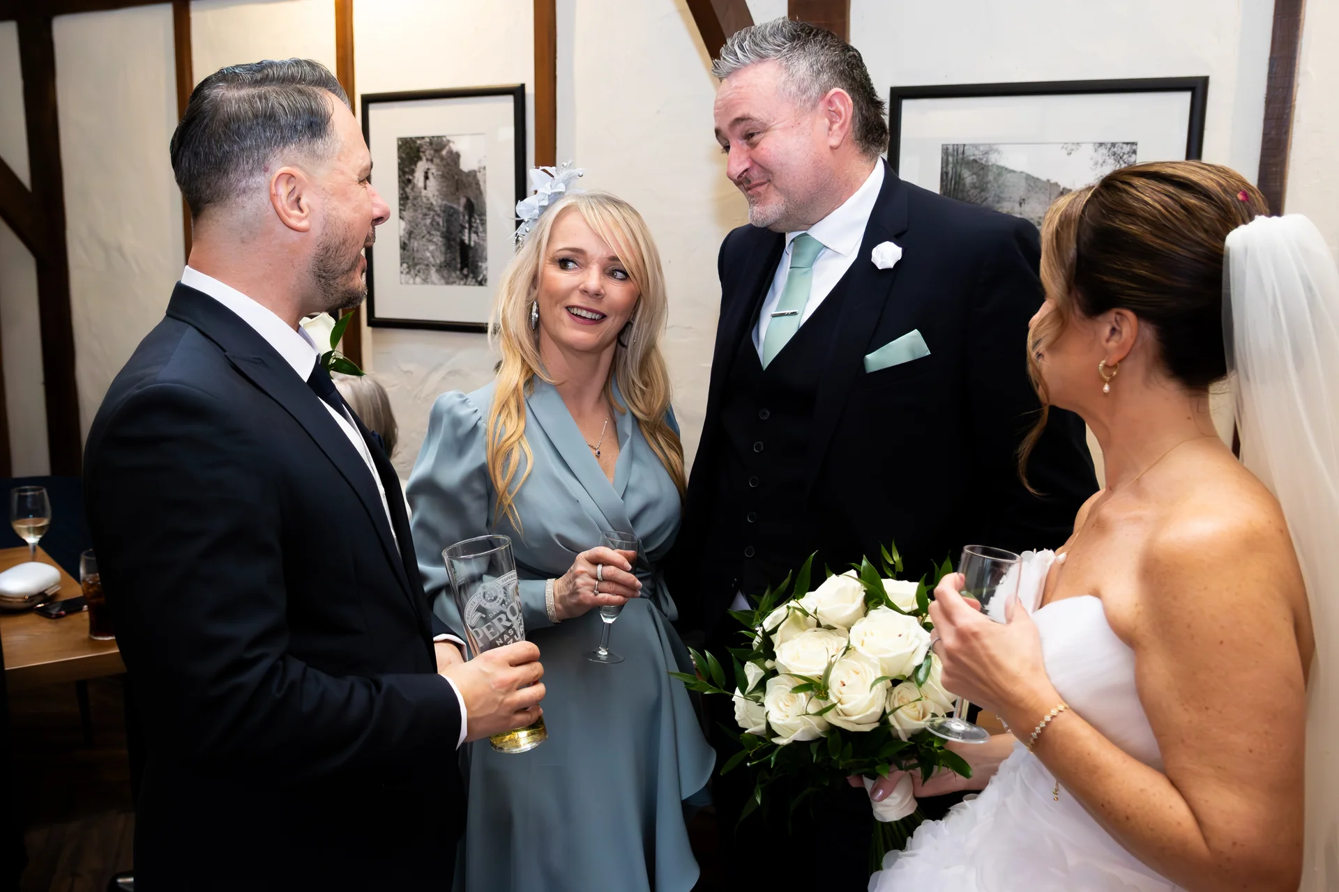 A bride in a white strapless gown and veil holds a white rose bouquet while chatting with guests at a wedding reception.